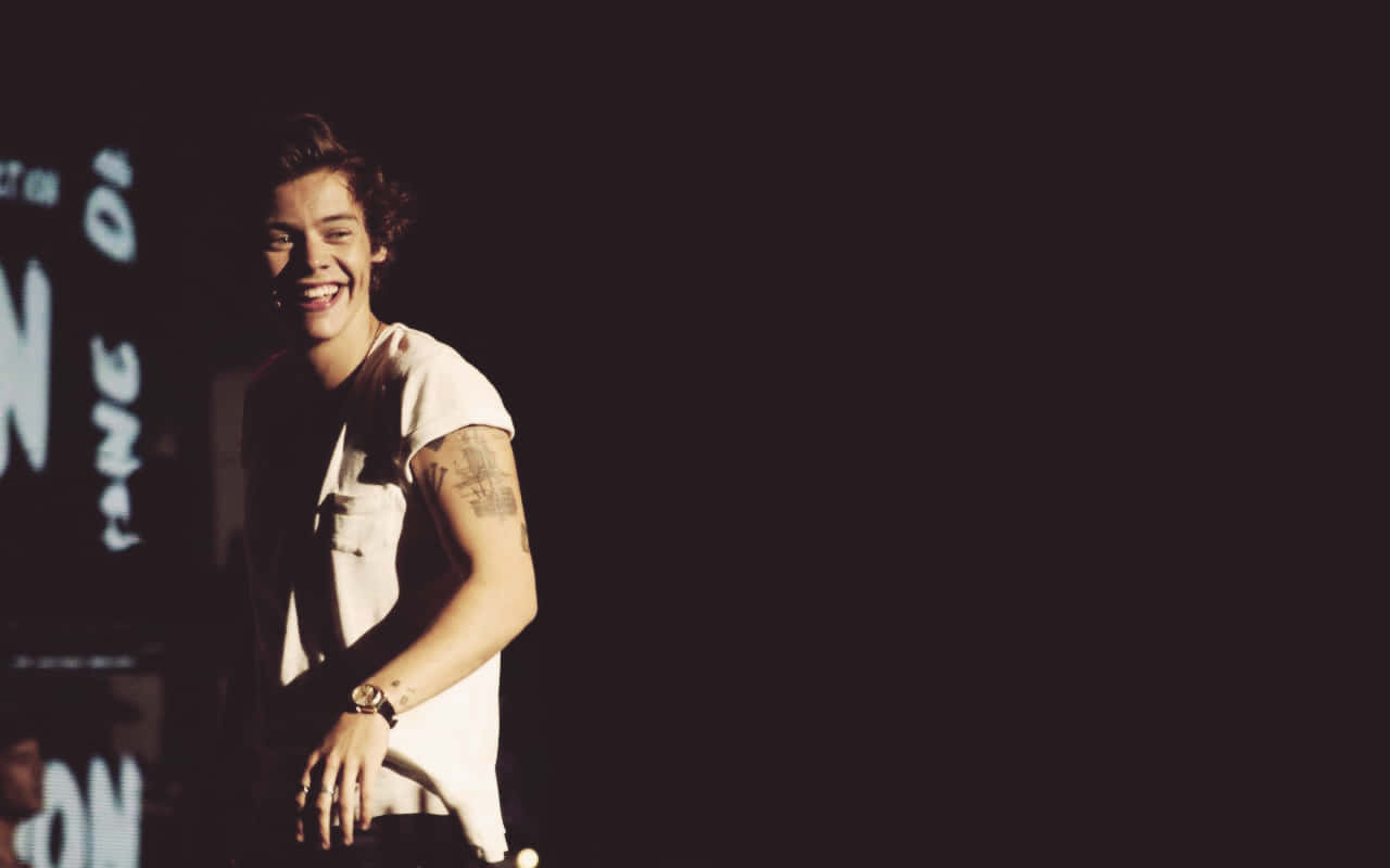 Harry Styles Laptop 桌布 壁紙 — 一位有刺青的年輕男子在舞台燈光下，對著黑暗背景中發光的文字燦爛大笑。