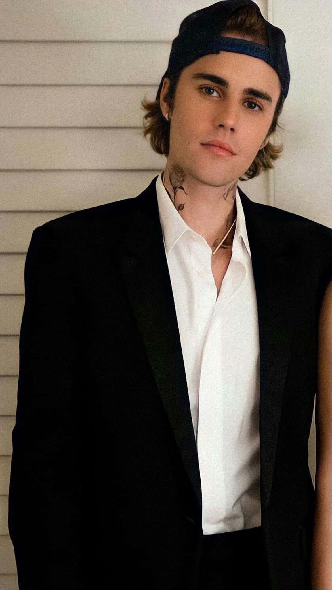 Justin Bieber 2021 桌布 壁紙 — 一位有刺青的年輕男子穿著黑色西裝外套和反戴的帽子，背景是白色鑲板牆。
