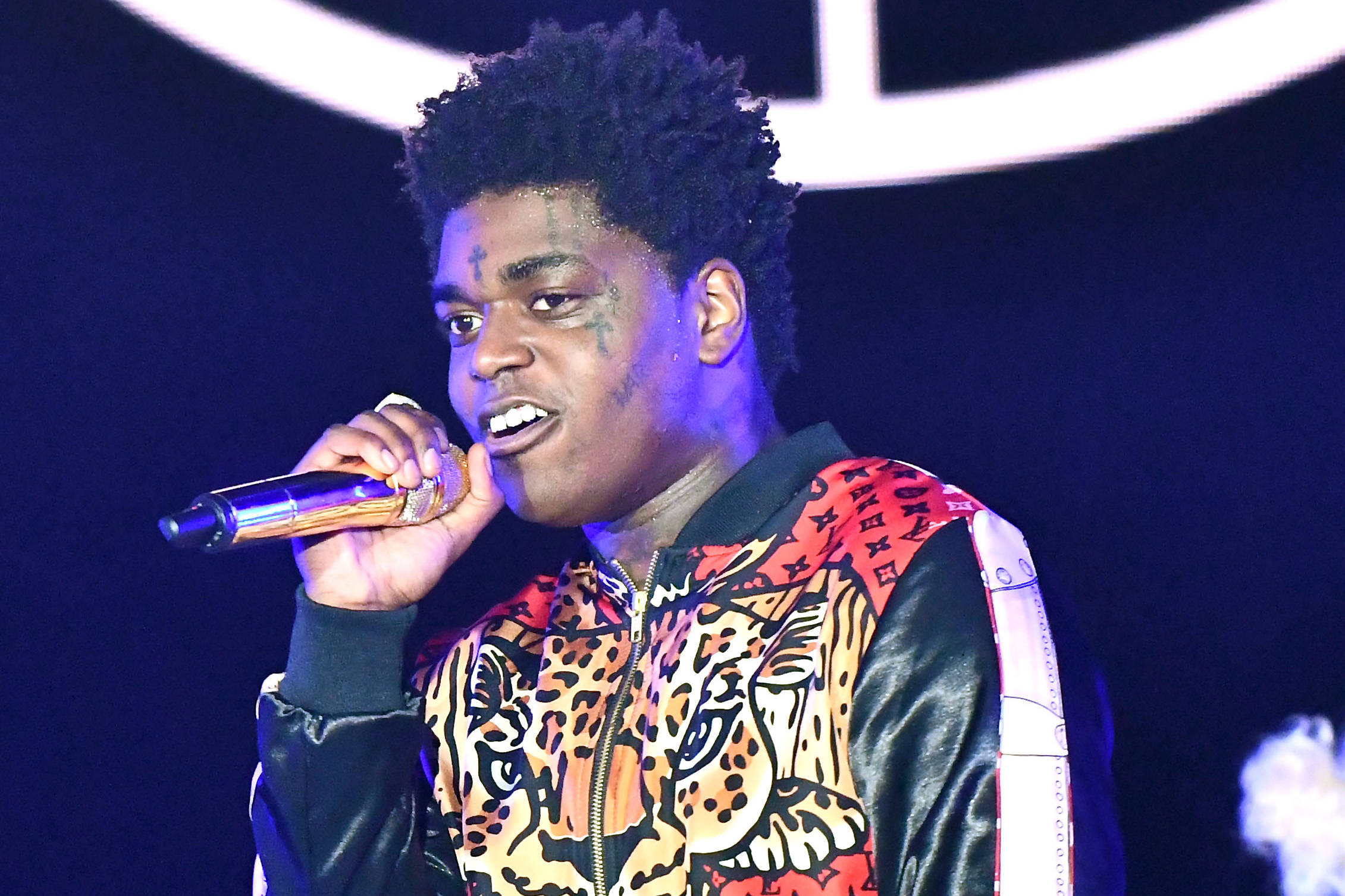 Kodak 桌布 壁紙 — 一位有刺青的年輕男表演者在舞台燈光下，穿著大膽的豹紋外套對著麥克風唱歌。