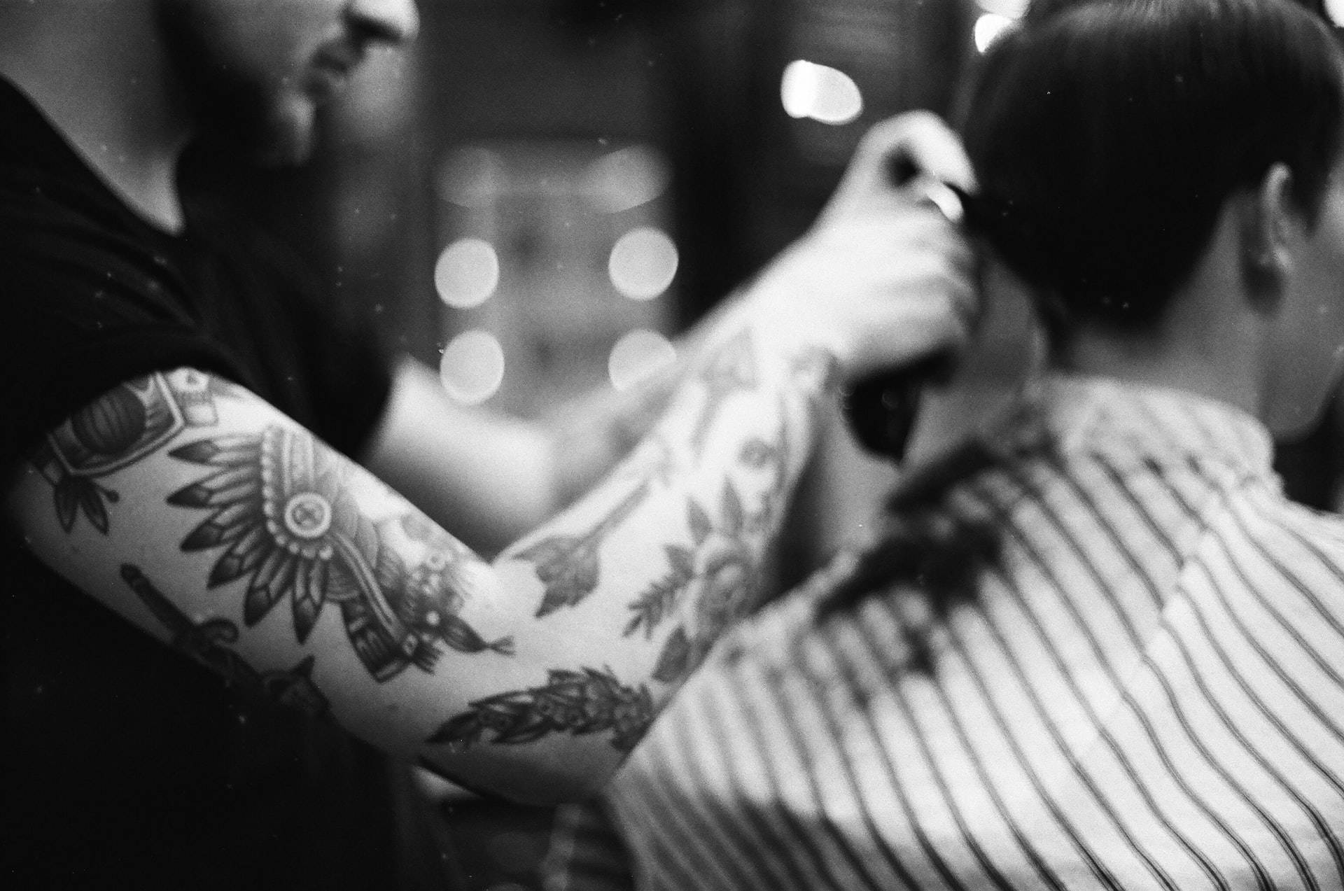 Tattoo 桌布 壁紙 — 一位有刺青的理髮師在光線昏暗的理髮店中，背景有散景燈光，細心地修剪顧客的頭髮。