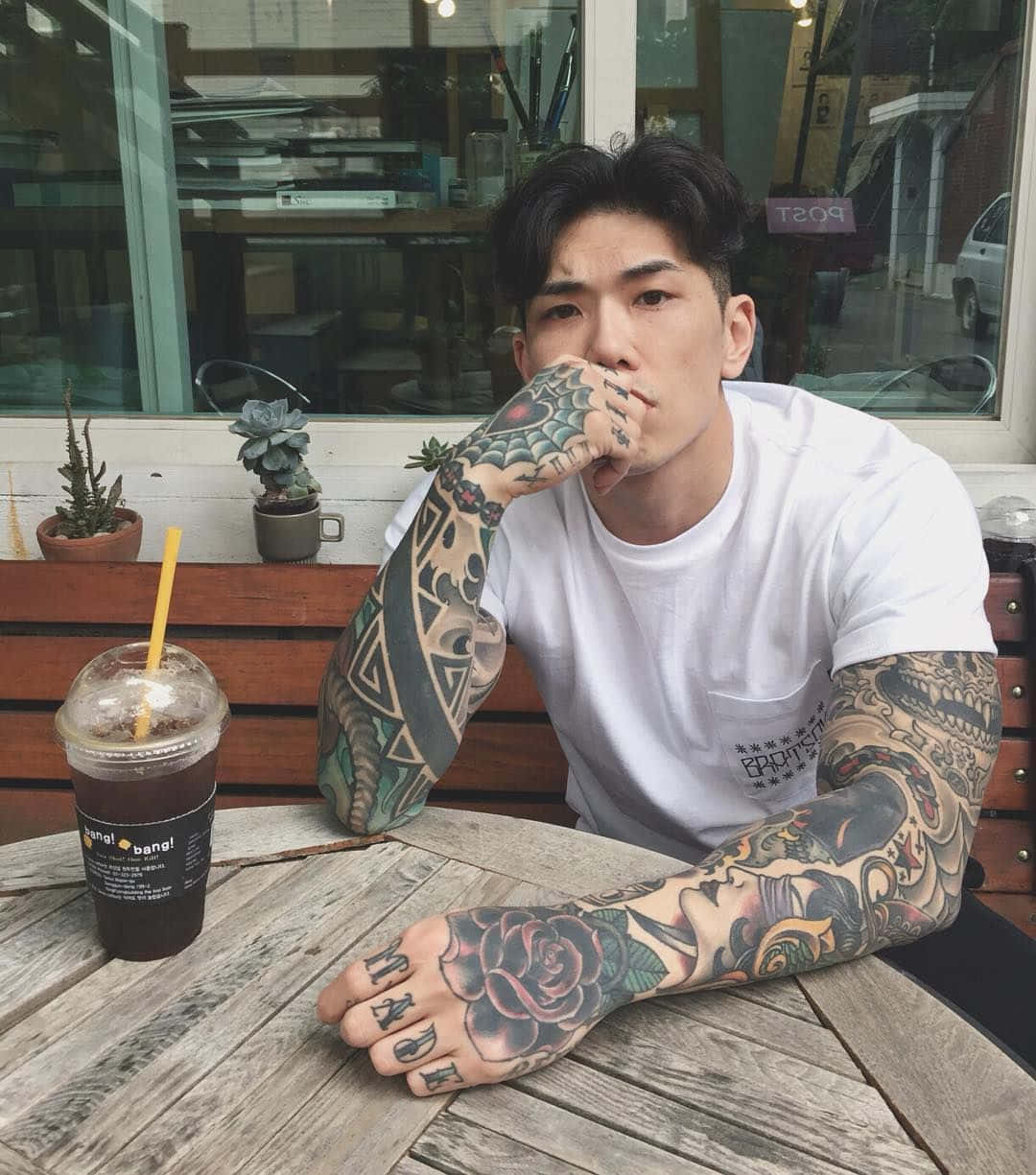 Tattoo Boy 桌布 壁紙 — 一位有刺青的男人穿著白色T恤坐在戶外的木桌旁，用吸管喝飲料。