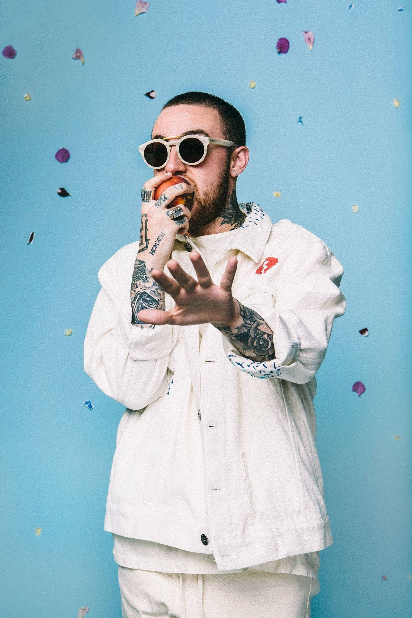 Mac Miller 桌布 壁紙 — 一位有刺青的男士，戴著白色太陽眼鏡和穿著白色外套，在藍色背景下吃柳橙，彩紙飄落。