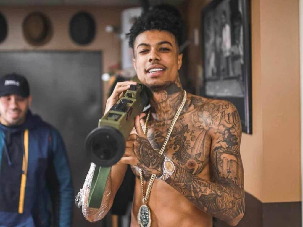 Celebrity 桌布 壁紙 — 一位有刺青的男士戴著金項鍊，在室內微笑著拿著一把大型綠色玩具槍。