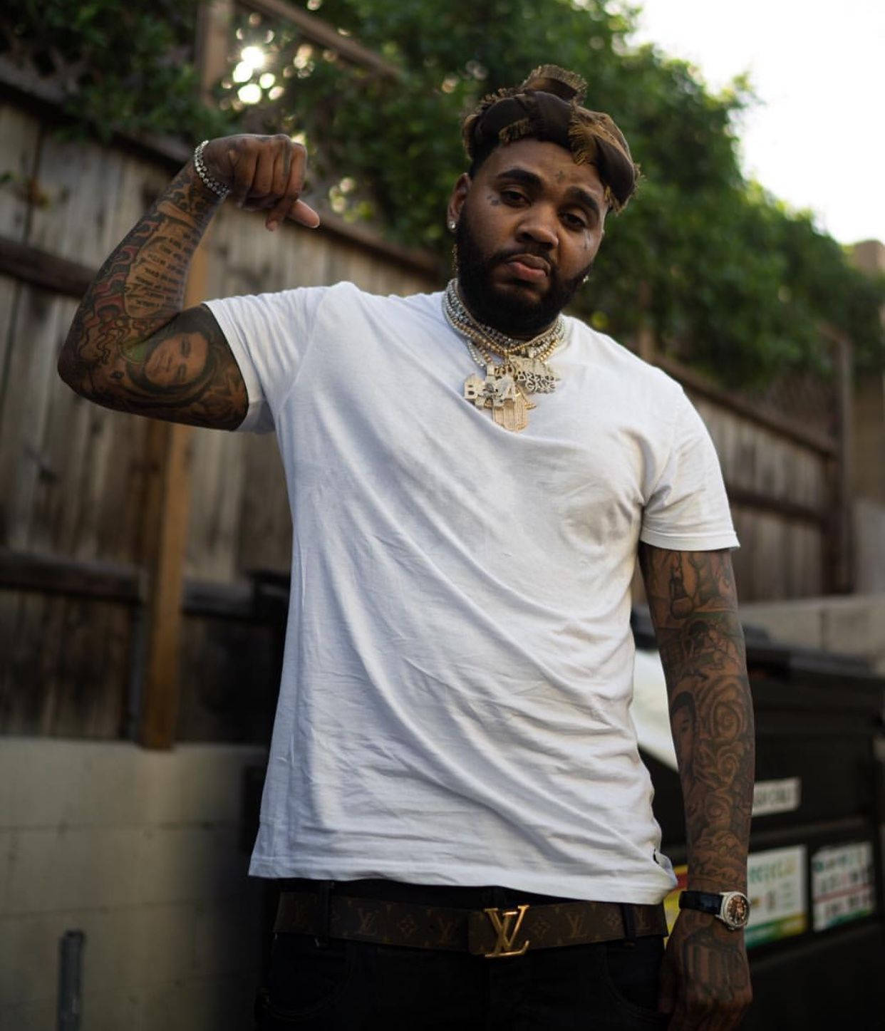 Kevin Gates 桌布 壁紙 — 一位有刺青的男士穿著白色T恤和戴著金鍊，在戶外木柵欄附近做出豎起拇指朝下的手勢。