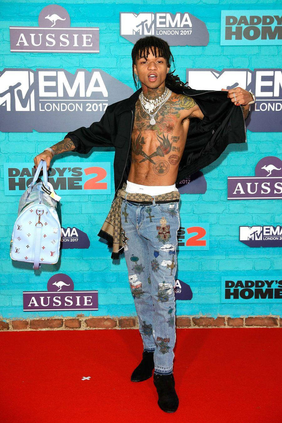 Swae Lee 桌布 壁紙 — 一位有刺青的男士穿著破洞牛仔褲和敞開的夾克，在藍色磚牆前的地毯上擺姿勢，牆上有活動標誌。