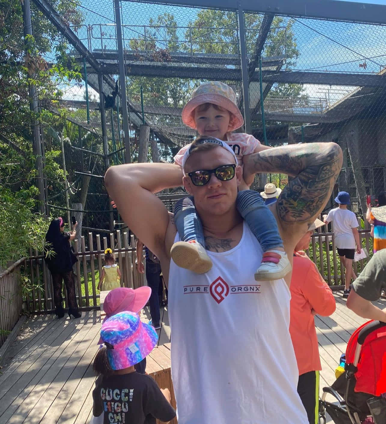 小孩動圖 桌布 壁紙 — 一位有刺青的男子在陽光明媚的動物園步道上，背著一個孩子，周圍是綠樹和遊客。