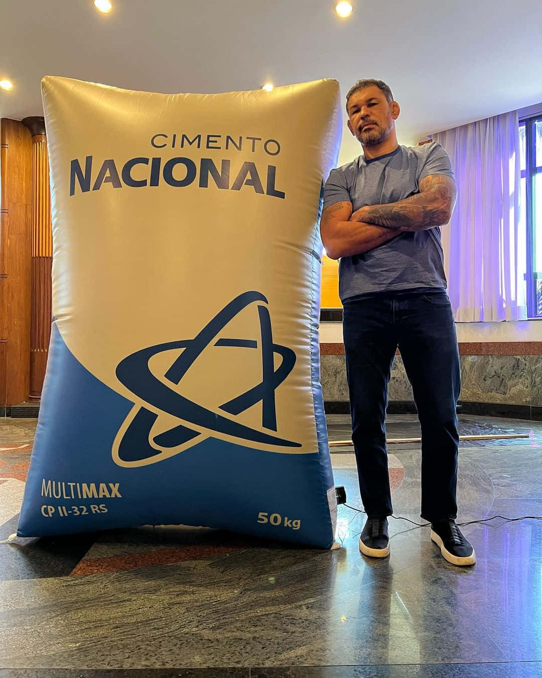 Celebrity 桌布 壁紙 — 一位有刺青的男子自信地站在一個標有「Cimento Nacional」的巨型充氣水泥袋旁，身處一個光潔的大廳。