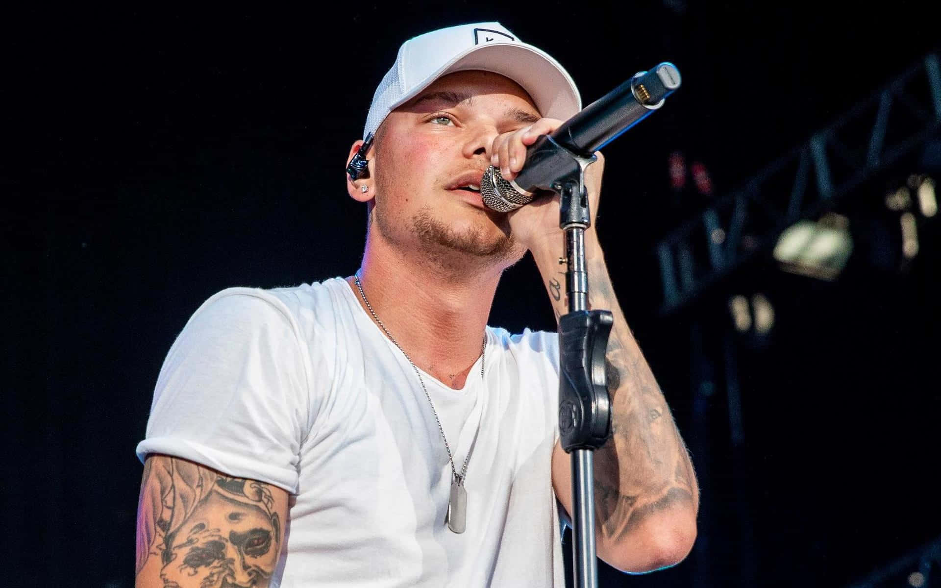 Kane Brown 桌布 壁紙 — 一位有刺青的男歌手，戴著白色帽子和T恤，在黑暗的舞台上熱情地對著麥克風演唱。