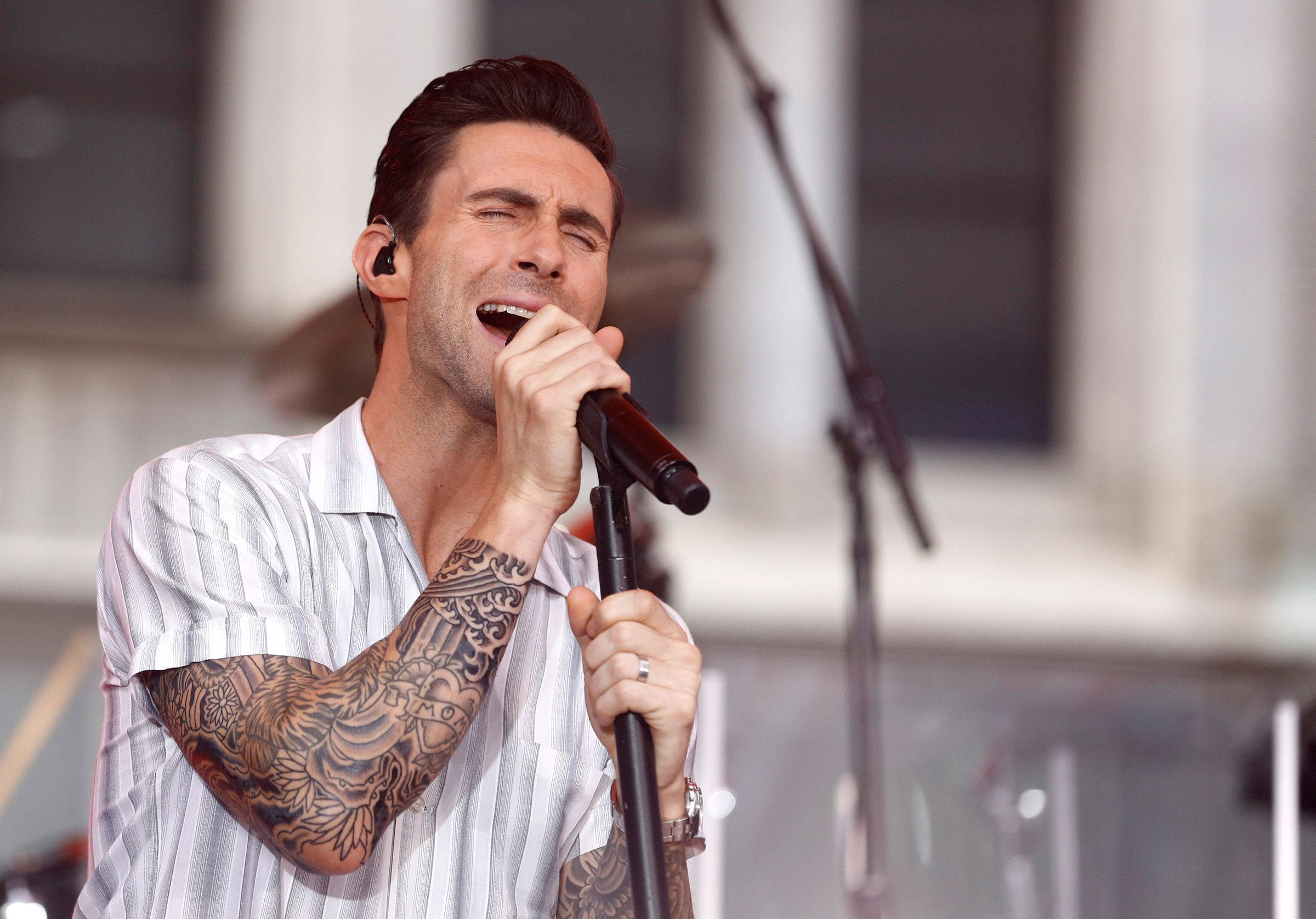Adam Levine 桌布 壁紙 — 一位有刺青的男歌手在明亮的舞台上熱情地對著麥克風表演。