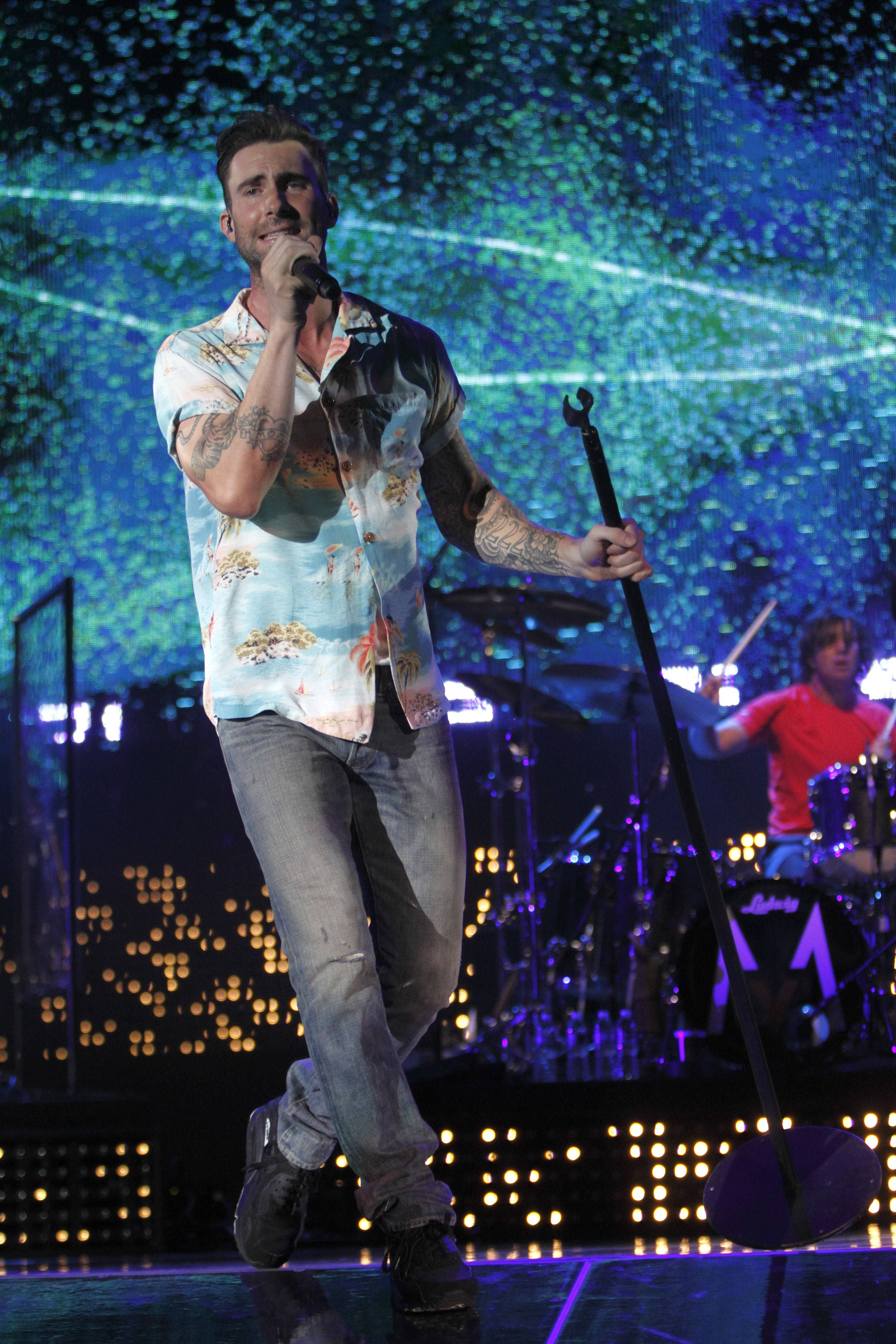 Adam Levine 桌布 壁紙 — 一位有刺青的男歌手在舞台上拿著麥克風，在充滿活力的藍色和綠色燈光下表演，背景有一位鼓手。
