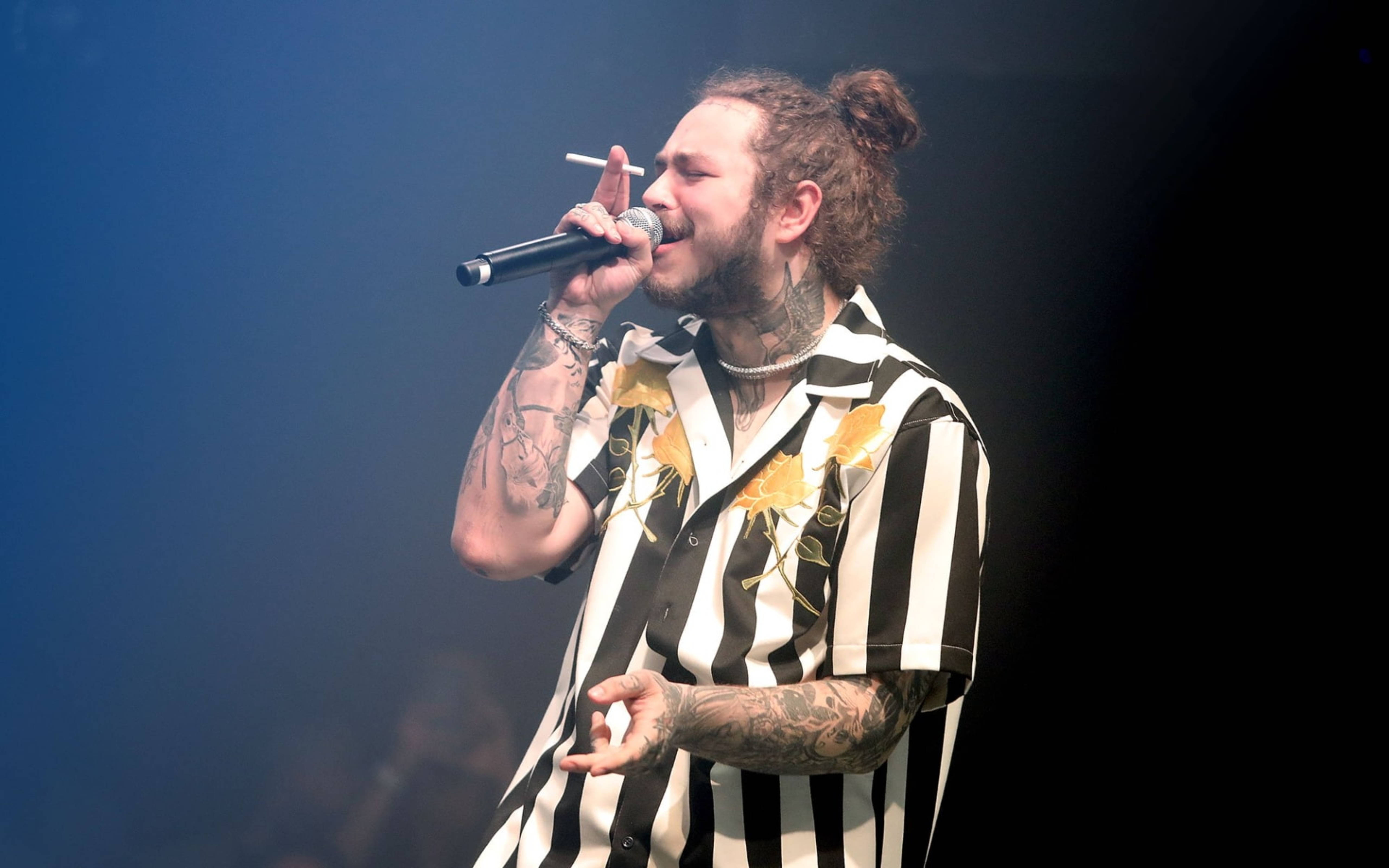 Post Malone 桌布 壁紙 — 一位有刺青的男歌手綁著髮髻，在藍色舞台燈光下拿著麥克風和香煙表演。