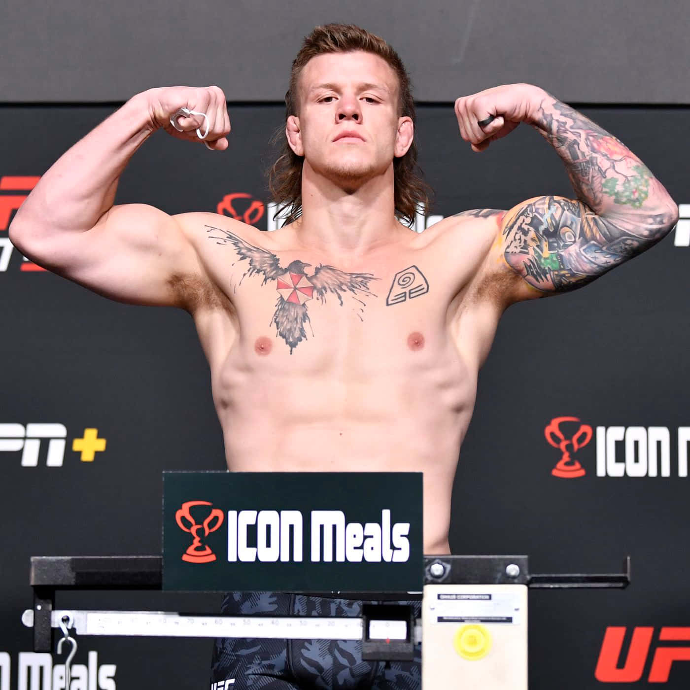 MMA 桌布 壁紙 — 一位有刺青的肌肉發達拳手在明亮的燈光下於 UFC 過磅活動中在磅秤上展示肌肉。