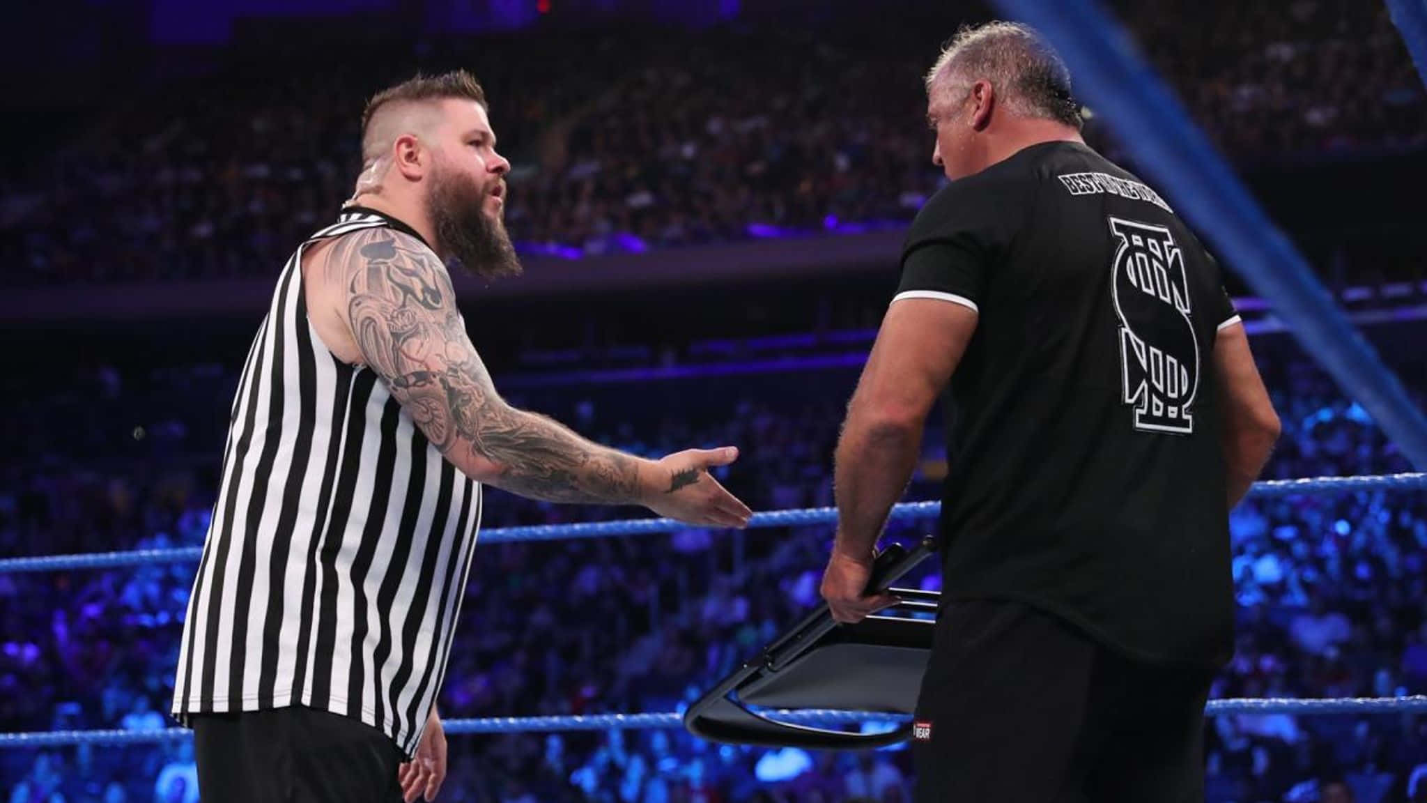 WWE 桌布 壁紙 — 一位有刺青的裁判穿著條紋衫，在燈光明亮的摔角場內與一名手持椅子的摔角手對峙。