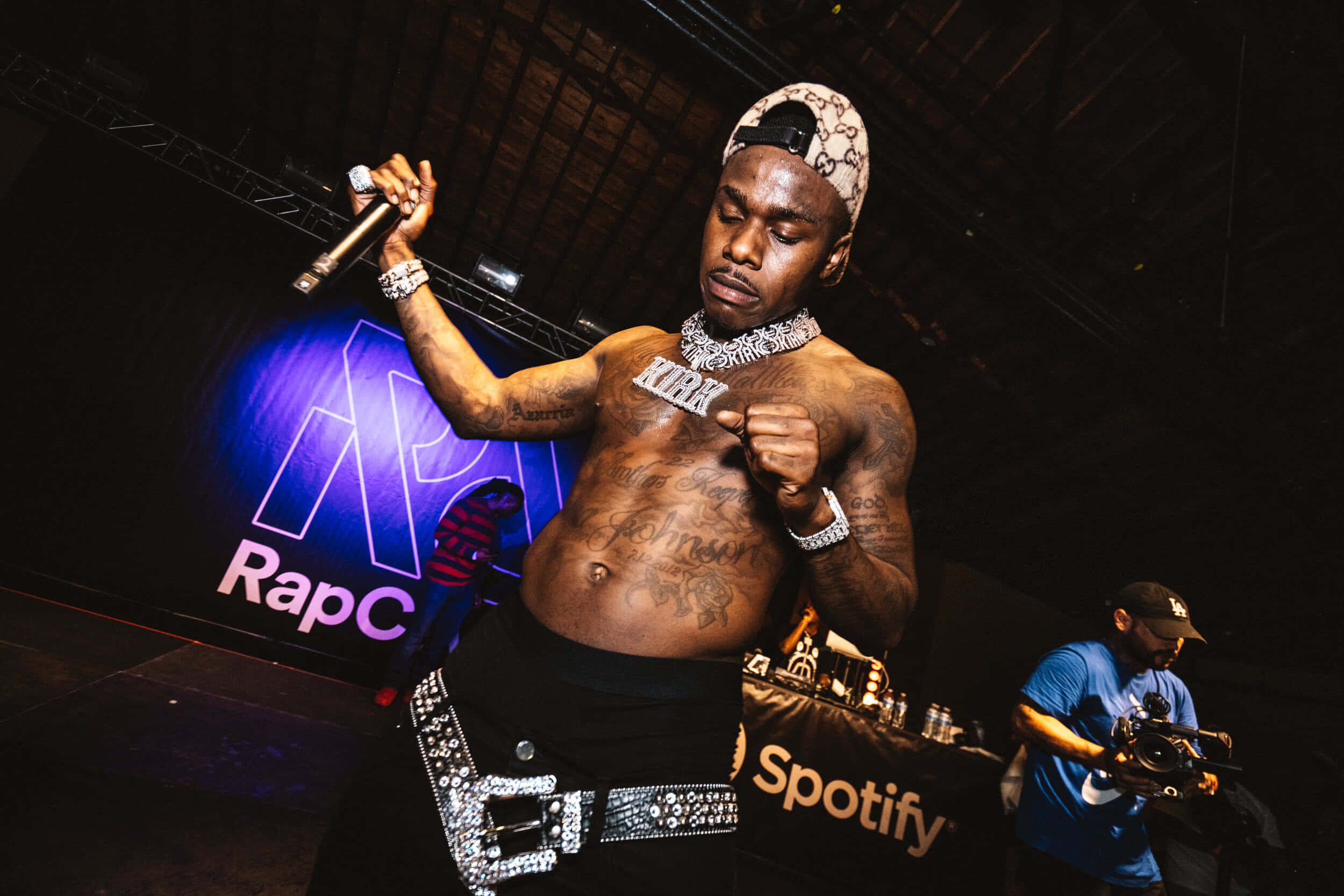 Dababy 桌布 壁紙 — 一位有刺青的說唱歌手在紫色聚光燈下拿著麥克風表演，身後有一位DJ。