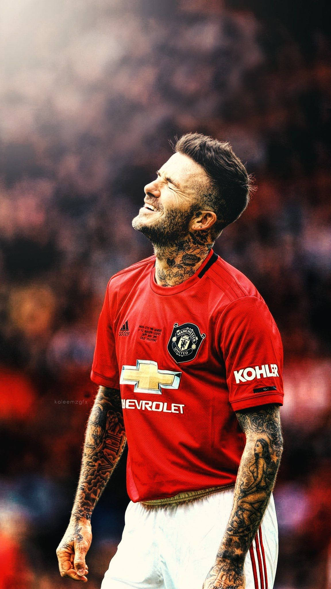 David Beckham 桌布 壁紙 — 一位有刺青的足球員穿著紅色曼聯球衣，在模糊的球場人群背景下閉著眼睛勝利地微笑。