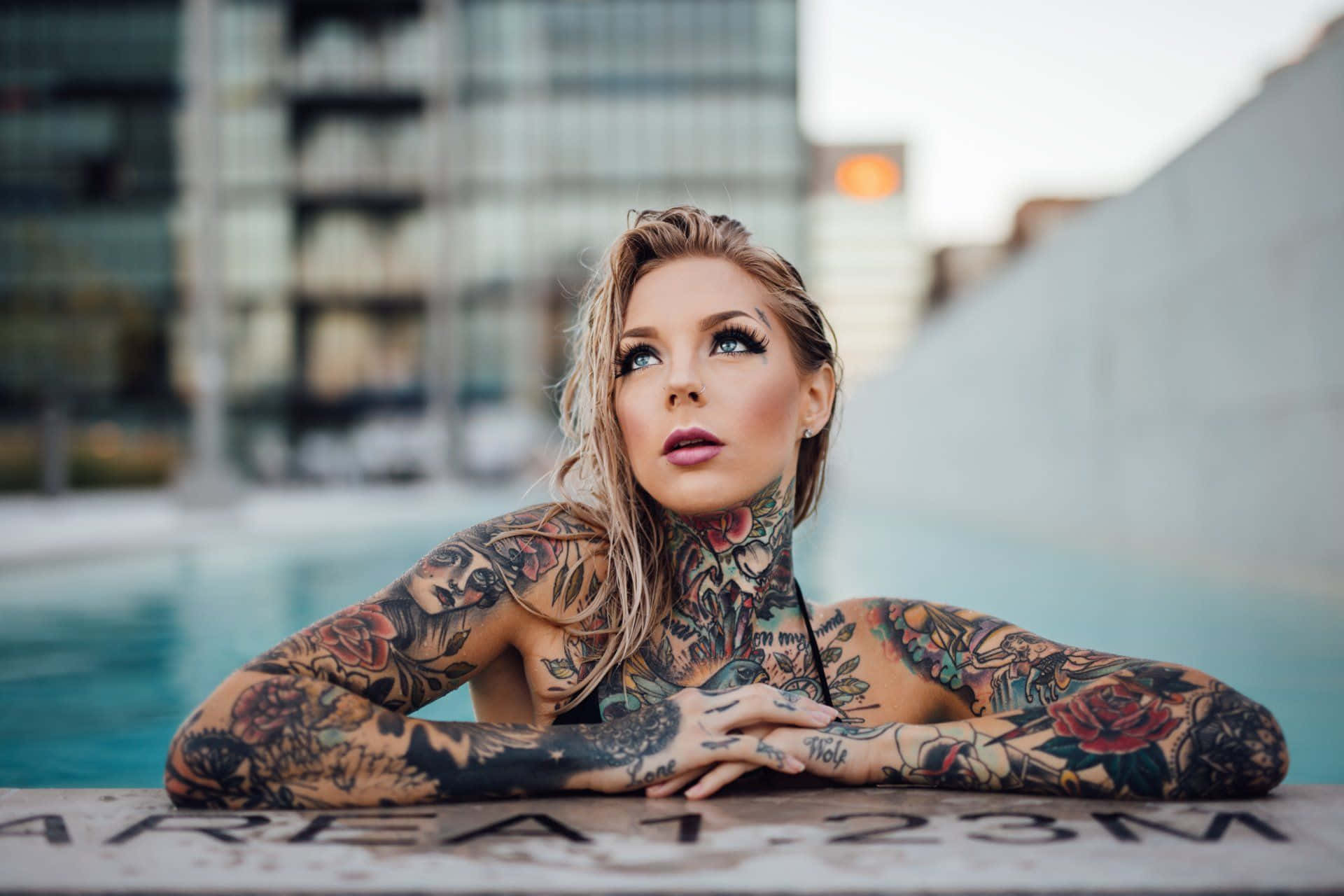 Tattoo 桌布 壁紙 — 一位有刺青的金髮女子將手臂擱在泳池邊，在暮色中對著模糊的城市背景向上凝視。