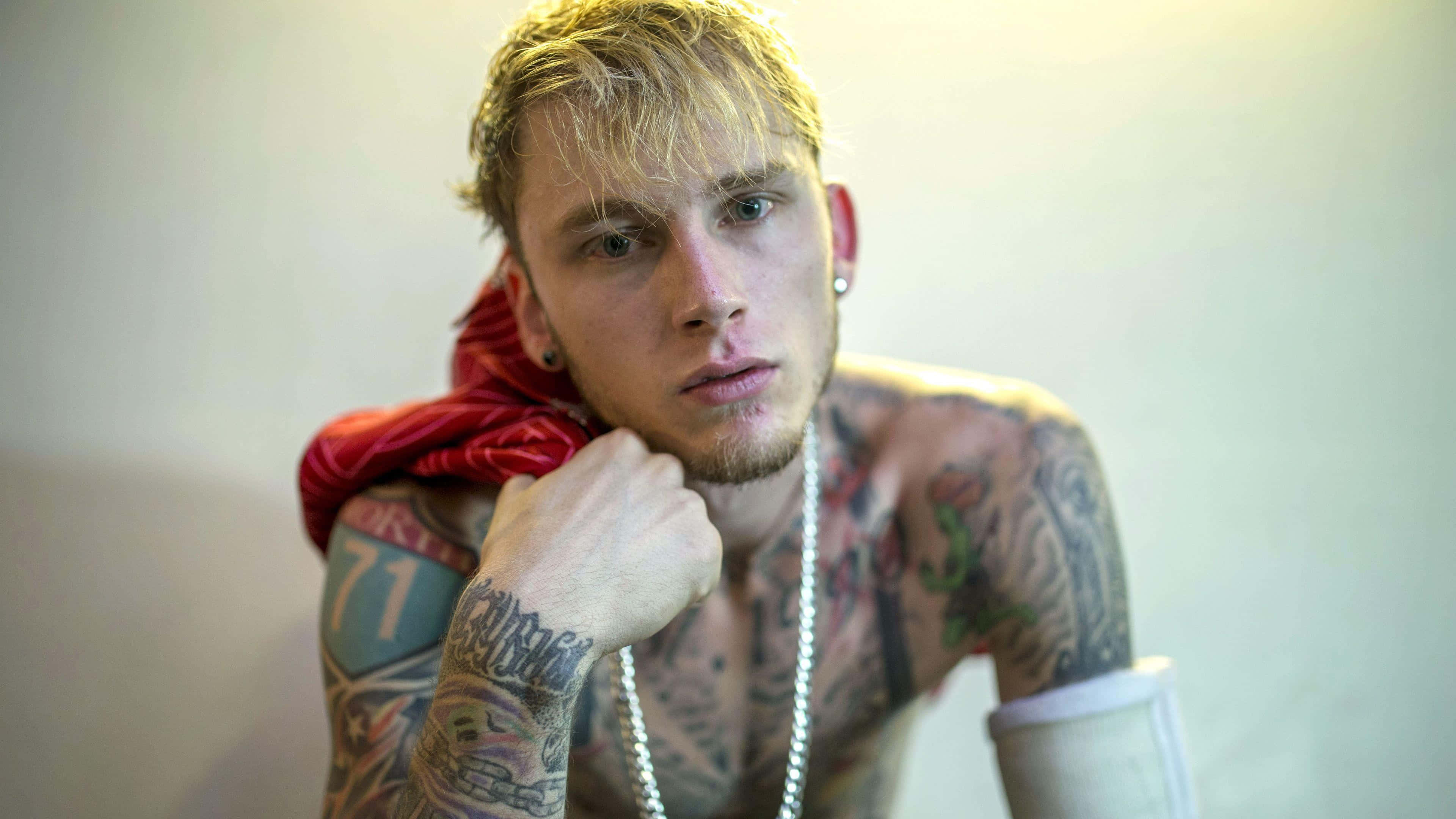 mgk 桌布 壁紙 — 一位有刺青的金髮年輕男子，戴著紅色圍巾，將下巴擱在拳頭上，靠著素牆。