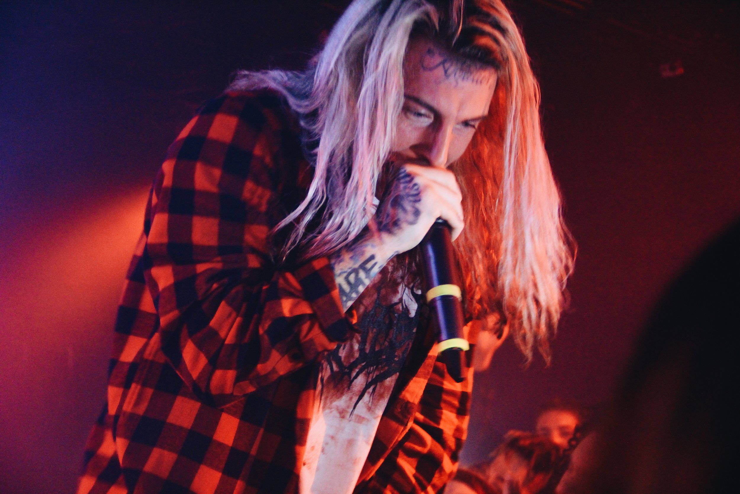 Ghostemane 桌布 壁紙 — 一位有刺青的音樂家，長長的金髮，在紅色舞台燈光下熱情地對著麥克風唱歌。