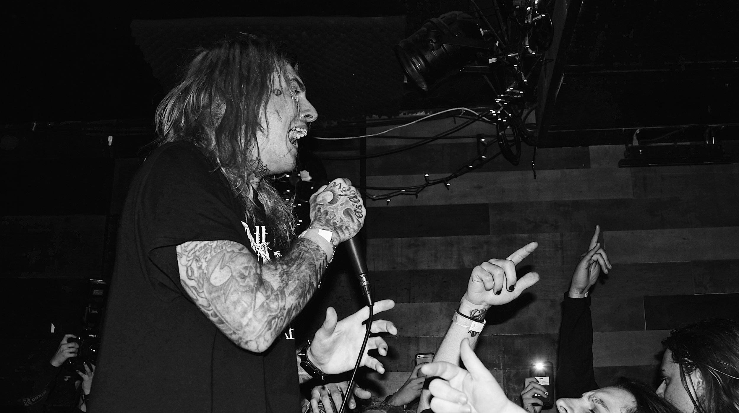 Ghostemane 桌布 壁紙 — 一位有刺青的音樂家在燈光昏暗的音樂會場地對著麥克風尖叫，粉絲們伸手向上。