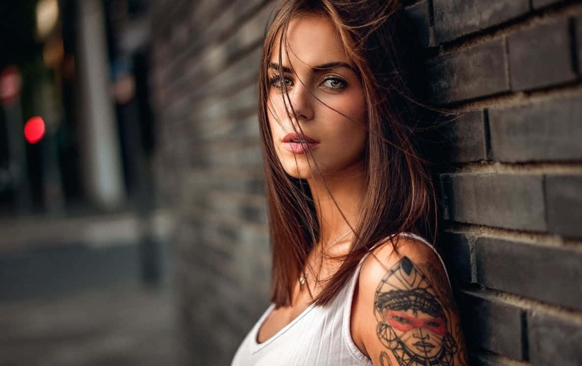 Halloween Tattoos 桌布 壁紙 — 一位有刺青肩膀的女性靠著深色磚牆，風吹動她的頭髮掠過臉龐。