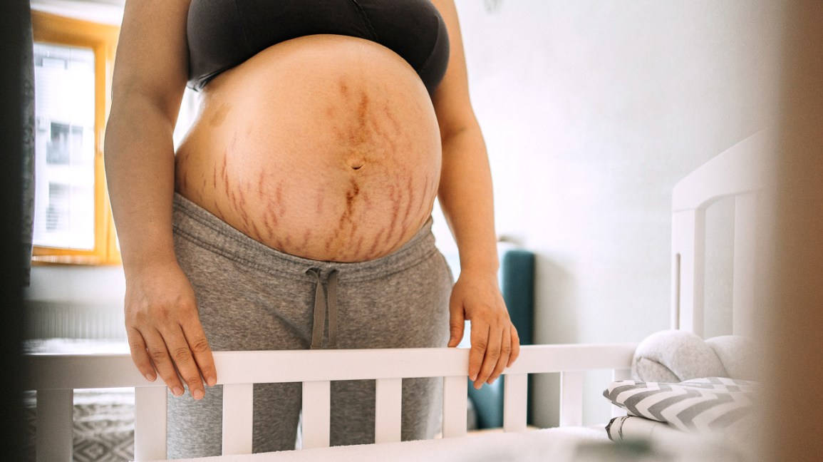 Pregnant 桌布 壁紙 — 一位有明顯妊娠紋的孕婦在柔和光線的臥室中俯身於白色嬰兒床上。