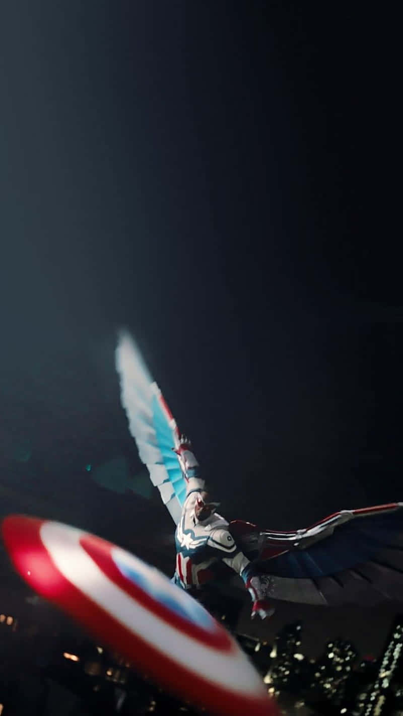 Marvel Falcon Logo 桌布 壁紙 — 一位有翅膀的超級英雄在夜間翱翔於城市上空，手持盾牌，下方燈火通明。