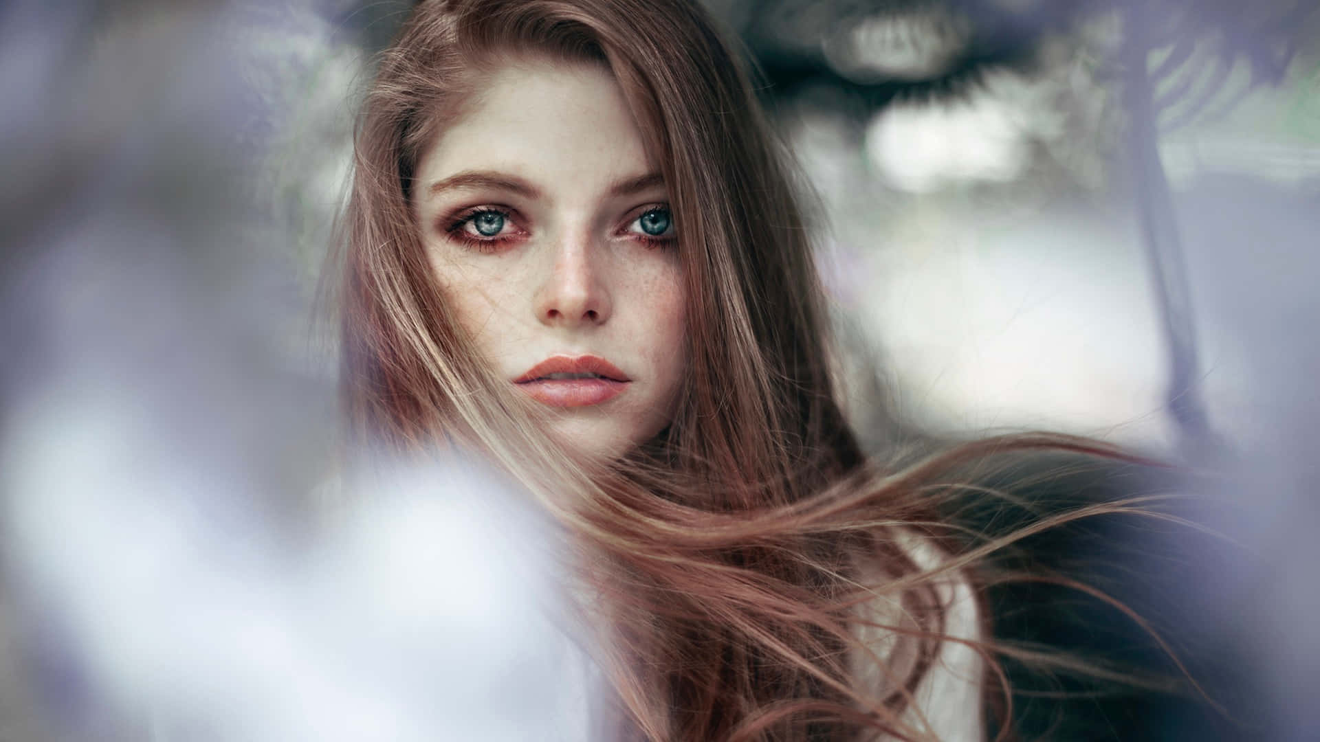 Beautiful Girl 桌布 壁紙 — 一位有雀斑和藍眼睛的女性在柔和自然光下透過模糊的樹葉凝視著。