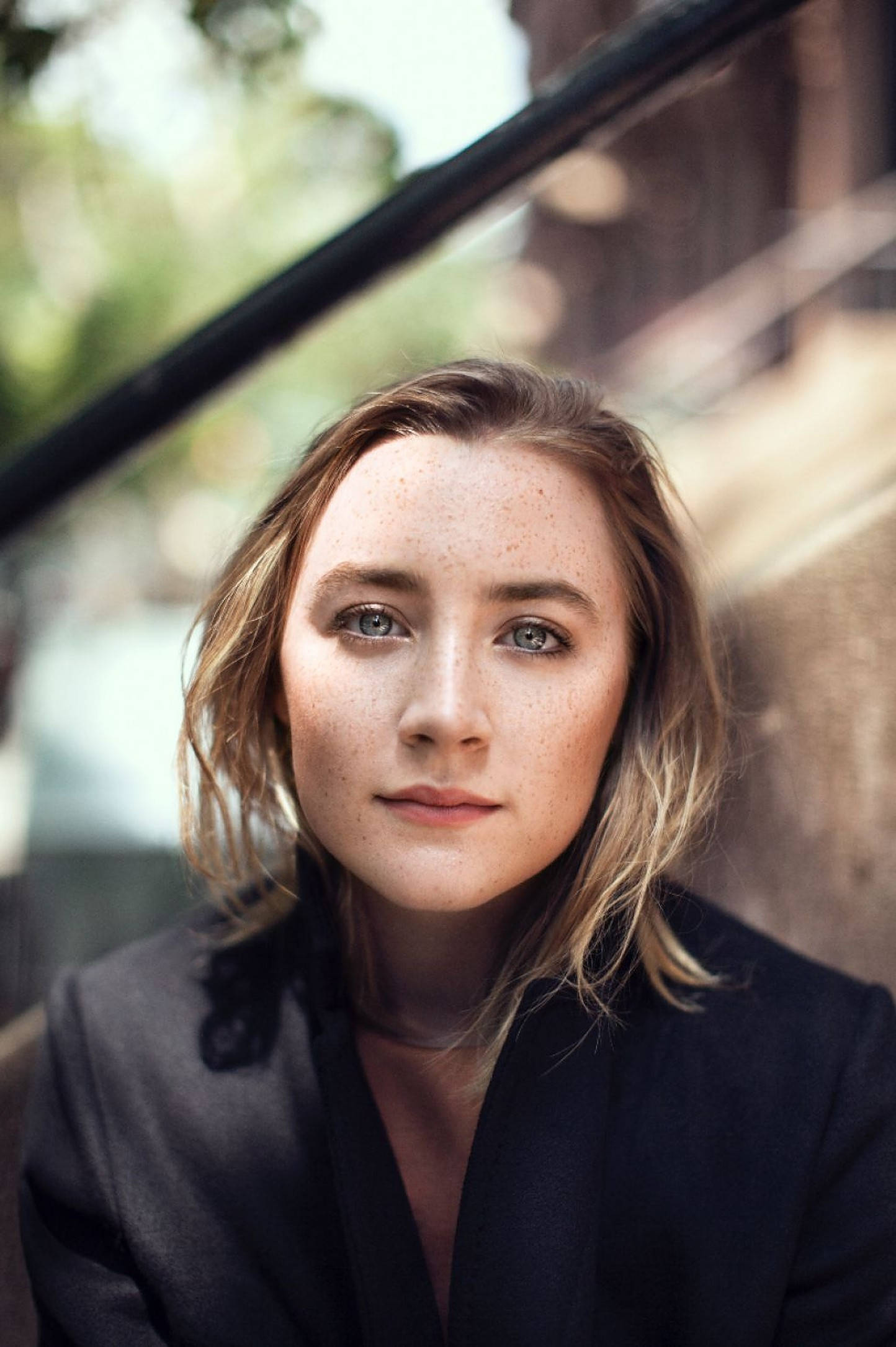Saoirse Ronan 桌布 壁紙 — 一位有雀斑和藍眼睛的年輕女性，平靜地看著鏡頭，坐在戶外欄杆附近。