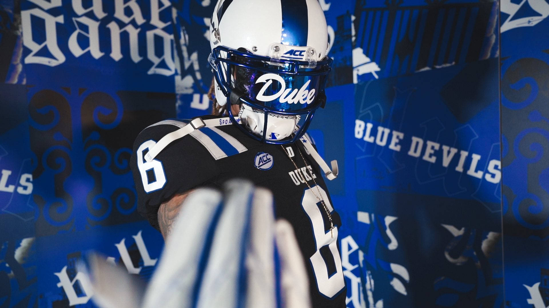 Duke Blue Devils 桌布 壁紙 — 一位杜克藍魔隊足球員身穿黑色和藍色制服，自信地站在品牌背景前。