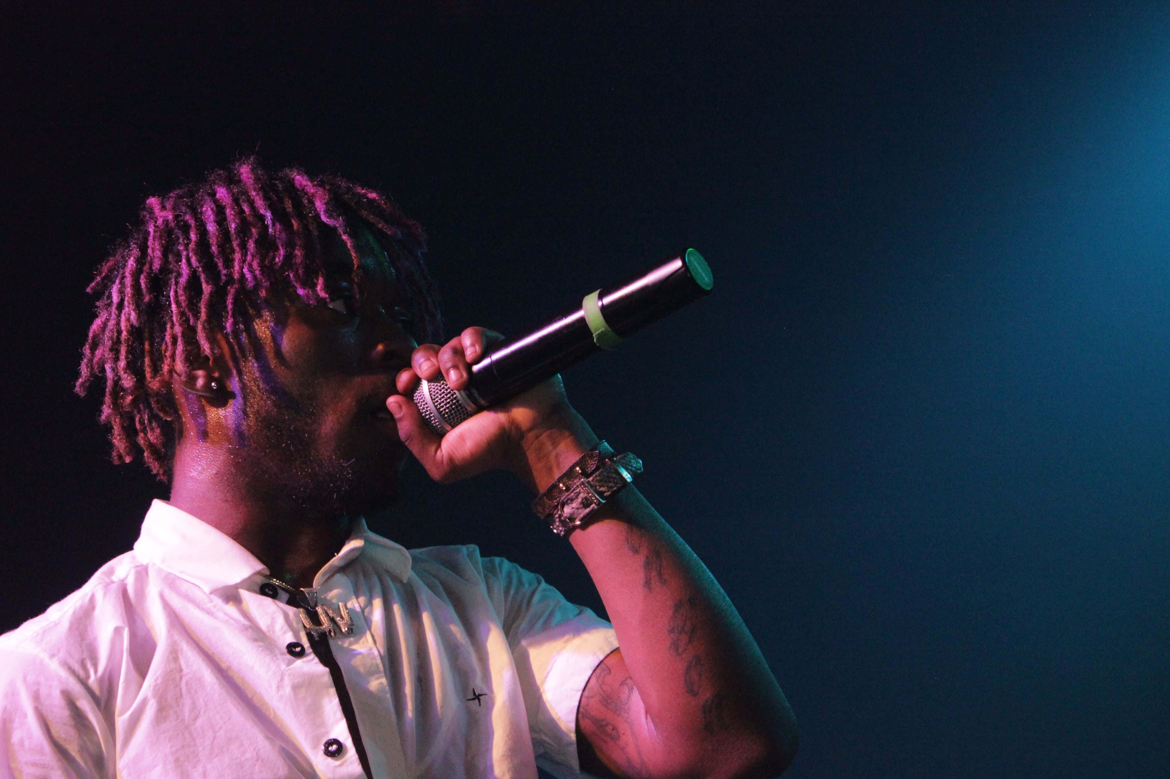 Lil Uzi Vert 桌布 壁紙 — 一位染有紫色辮子的饒舌歌手，身穿白襯衫，在藍色舞台燈光下對著麥克風唱歌。