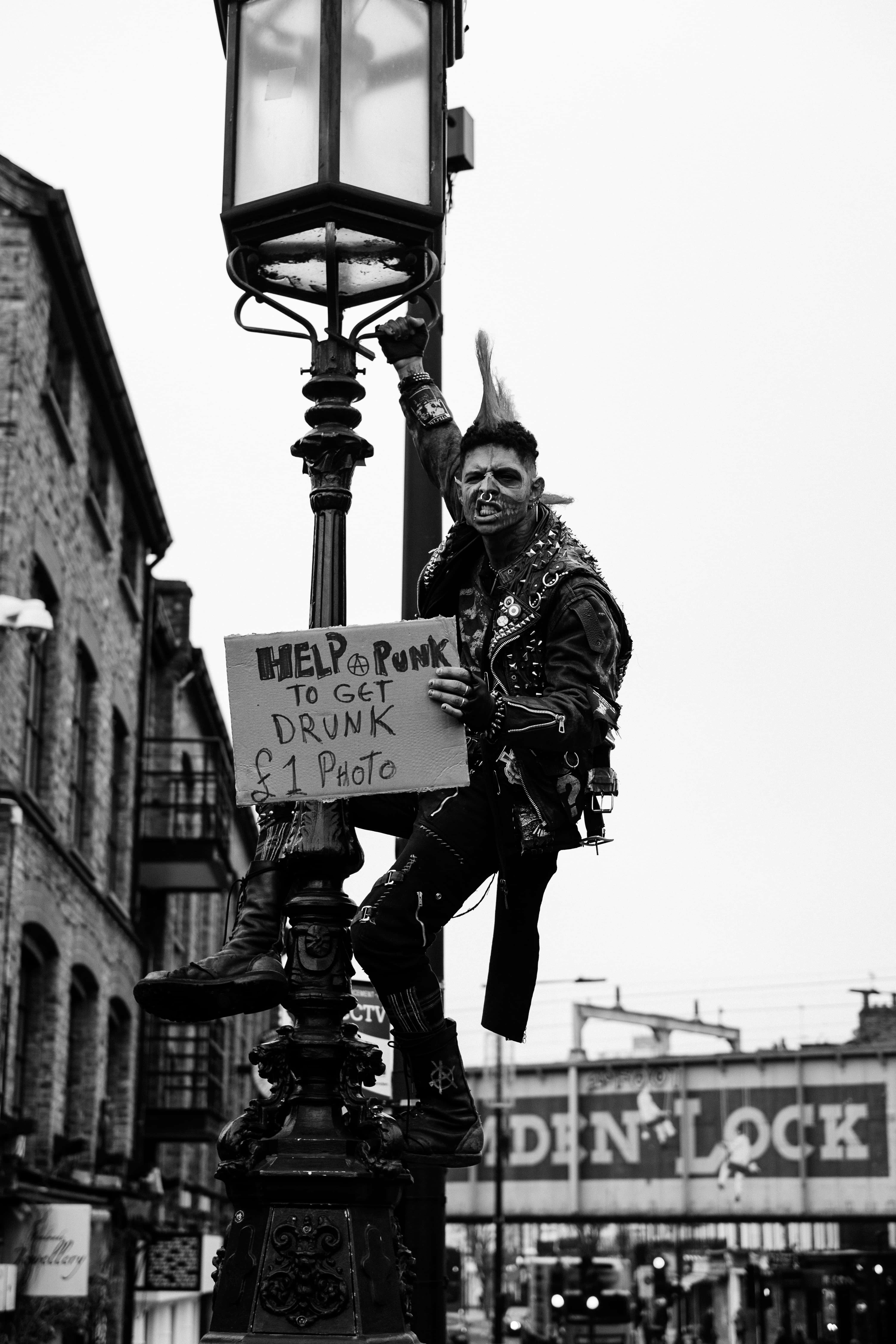 punk 桌布 壁紙 — 一位梳著尖刺頭髮、臉上塗著顏料的龐克搖滾歌手，攀爬在一盞復古路燈上，舉著一個牌子，在骯髒的城市環境中提供拍照賺錢的服務。