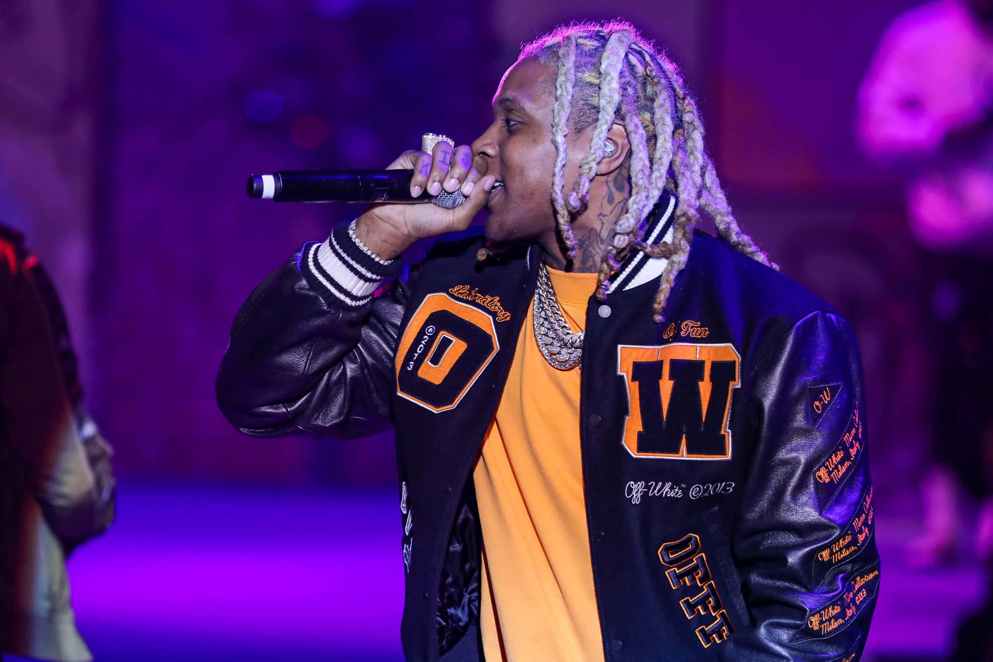 Lil Durk 桌布 壁紙 — 一位梳著辮子的饒舌歌手在紫色燈光下穿著帶有橙色字母的黑色校隊夾克在舞台上表演。