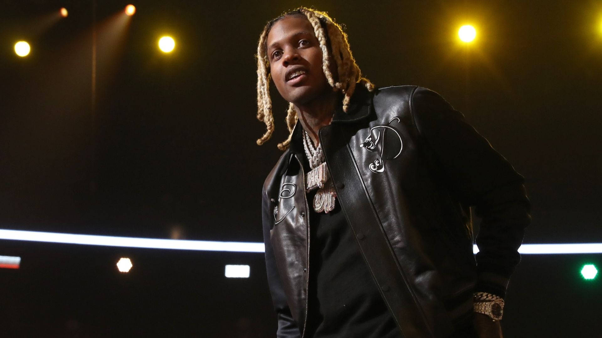 Lil Durk 桌布 壁紙 — 一位梳著辮子的饒舌歌手自信地站在昏暗的舞台上，在溫暖的聚光燈下，身穿黑色皮夾克和戴著金鍊。