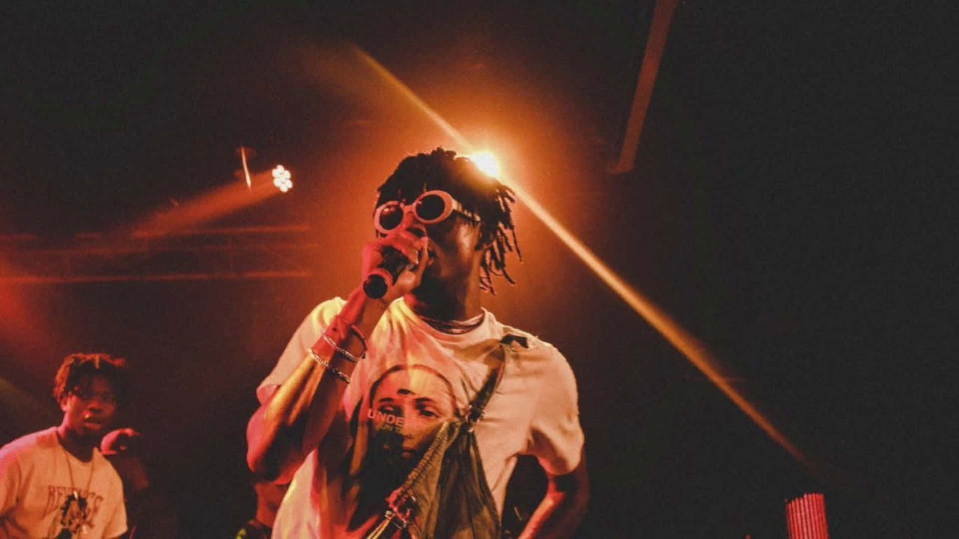 Playboi Carti 桌布 壁紙 — 一位梳著辮子頭、戴圓框眼鏡的饒舌歌手，在溫暖的舞台燈光下對著麥克風熱情表演。