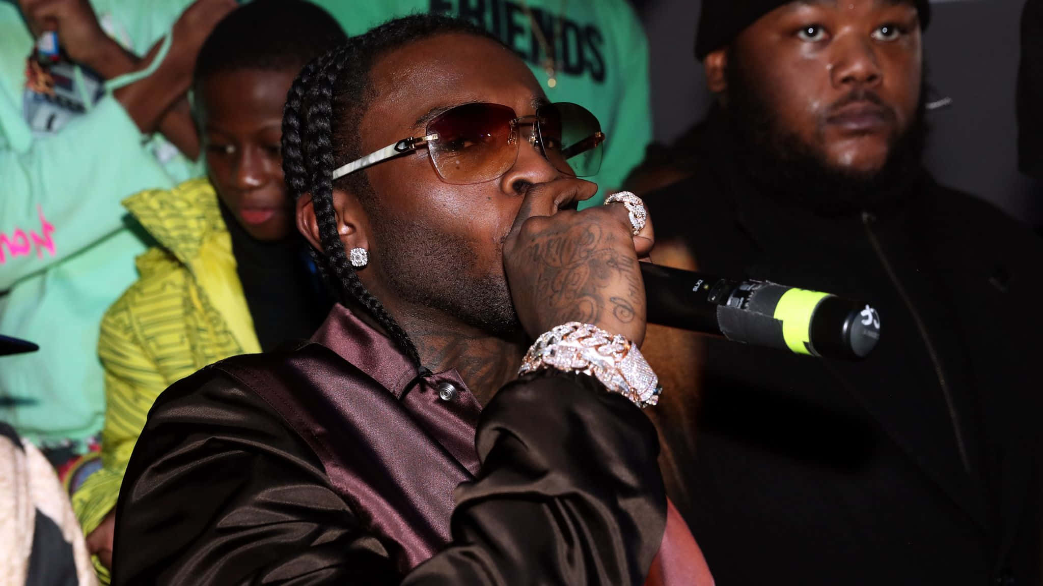 Dead Rappers 桌布 壁紙 — 一位梳著辮子頭、戴著太陽眼鏡的饒舌歌手，穿著閃亮的棕色襯衫和鑽石首飾，對著麥克風表演。