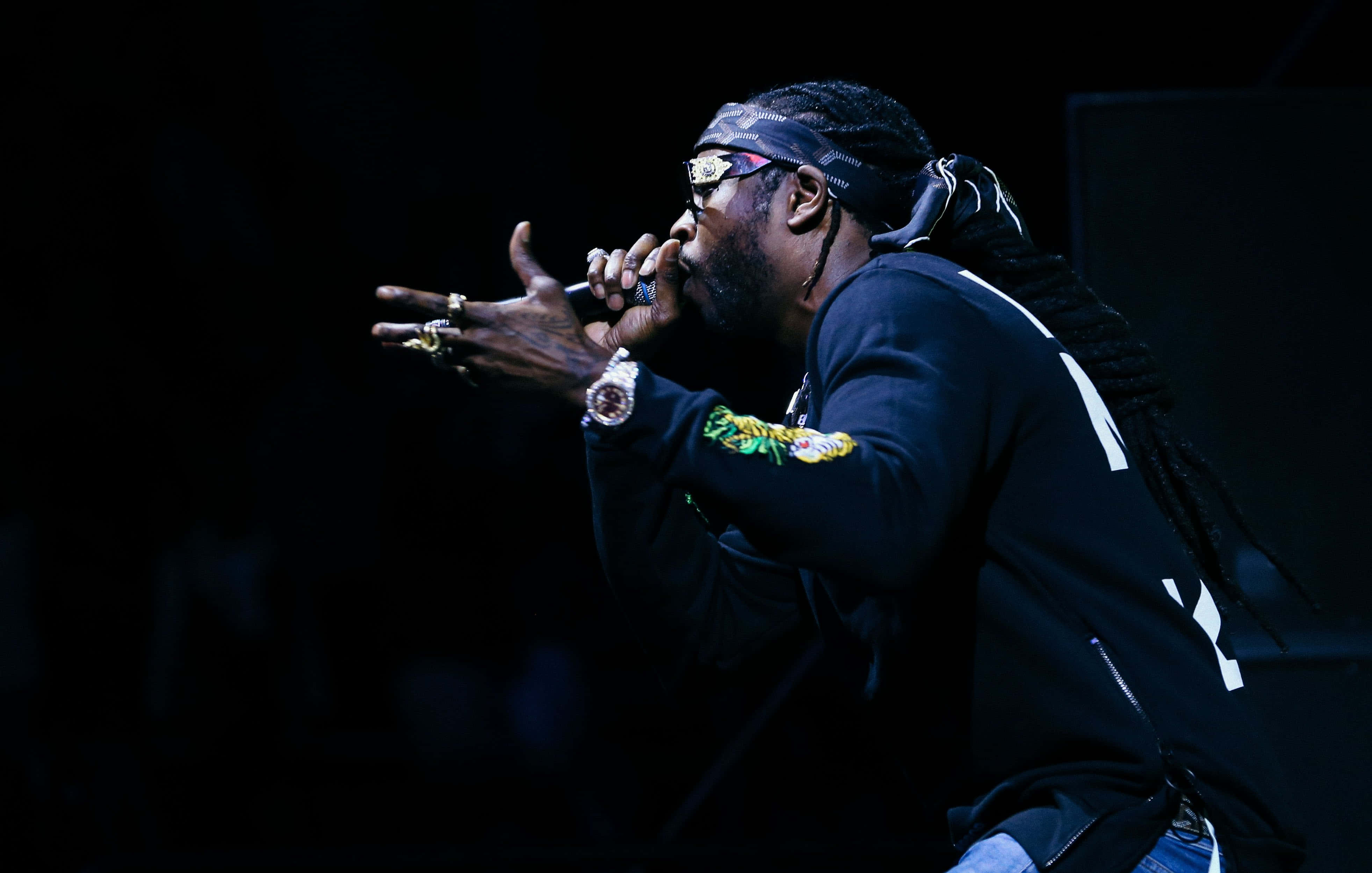 2 Chainz 桌布 壁紙 — 一位梳著辮子頭、戴著太陽眼鏡的饒舌歌手，在戲劇性的舞台燈光下，對著麥克風充滿激情地表演。