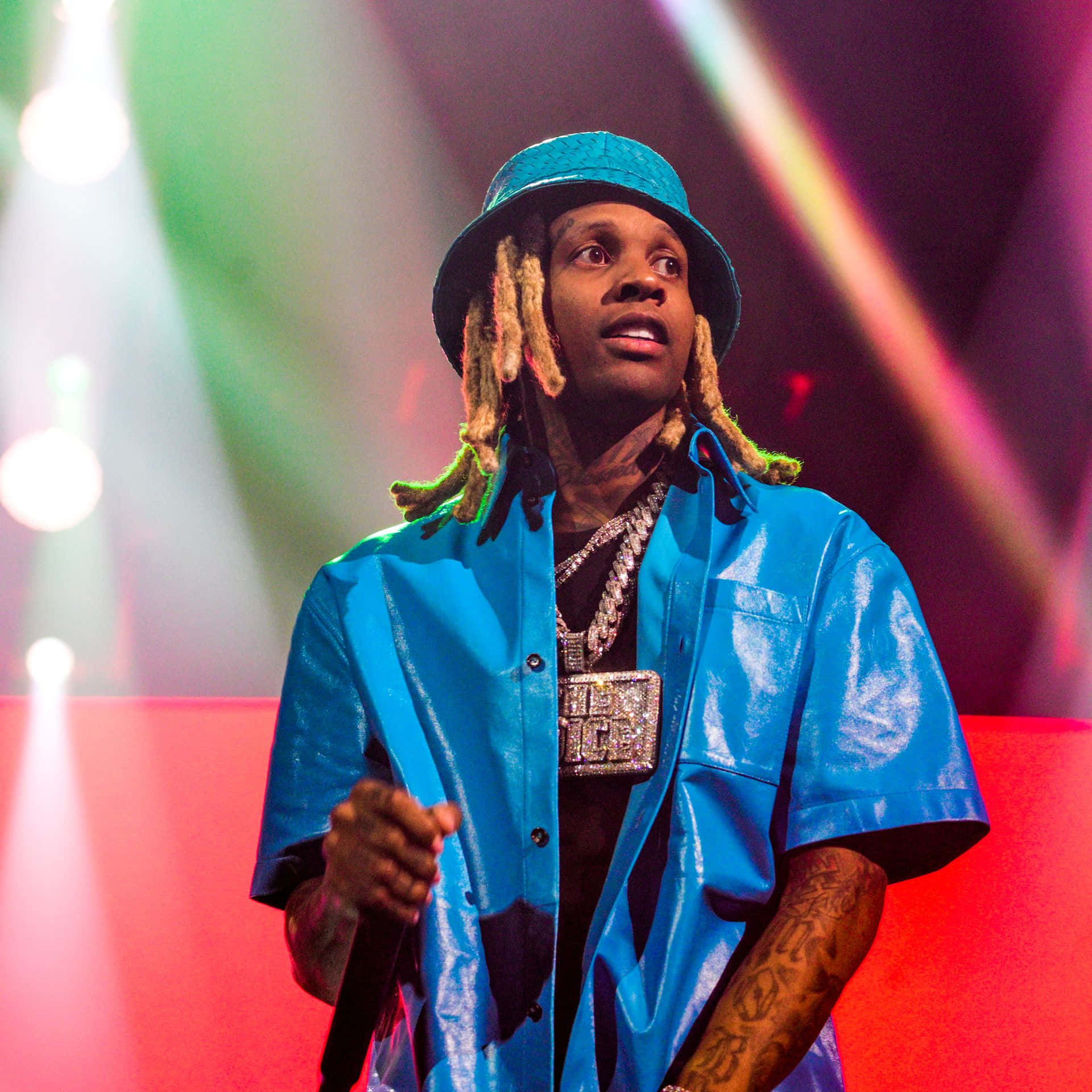 Lil Durk 桌布 壁紙 — 一位梳著辮子頭、戴藍色帽子的饒舌歌手在彩色聚光燈下表演，身穿閃亮藍色襯衫和厚重珠寶。