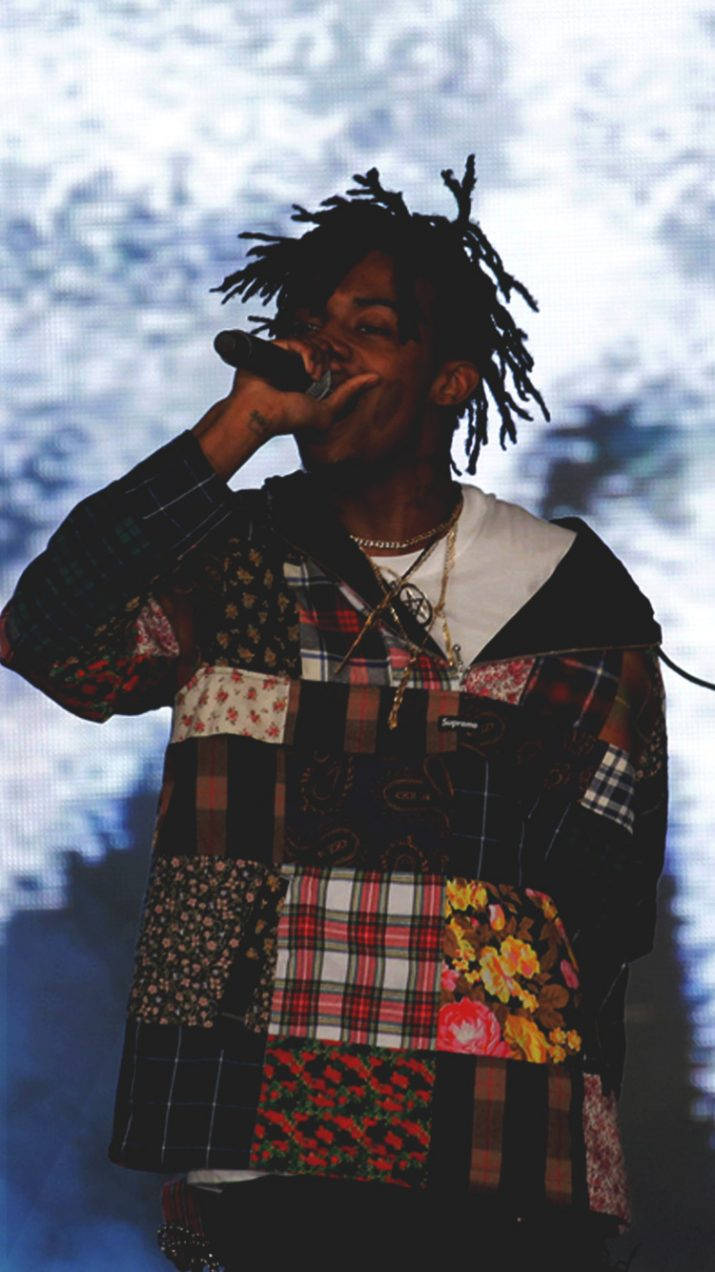 Carti 桌布 壁紙 — 一位梳著辮子頭的饒舌歌手，身穿色彩繽紛的拼布外套，在明亮的舞台燈光下對著麥克風唱歌。