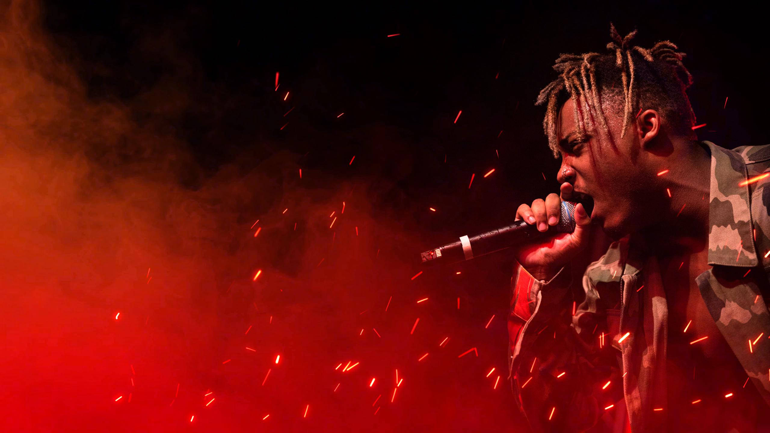 Juice Wrld Desktop 桌布 壁紙 — 一位梳著辮子頭的饒舌歌手熱情地對著麥克風表演，紅色火花在煙霧繚繞的舞台燈光中飛舞。