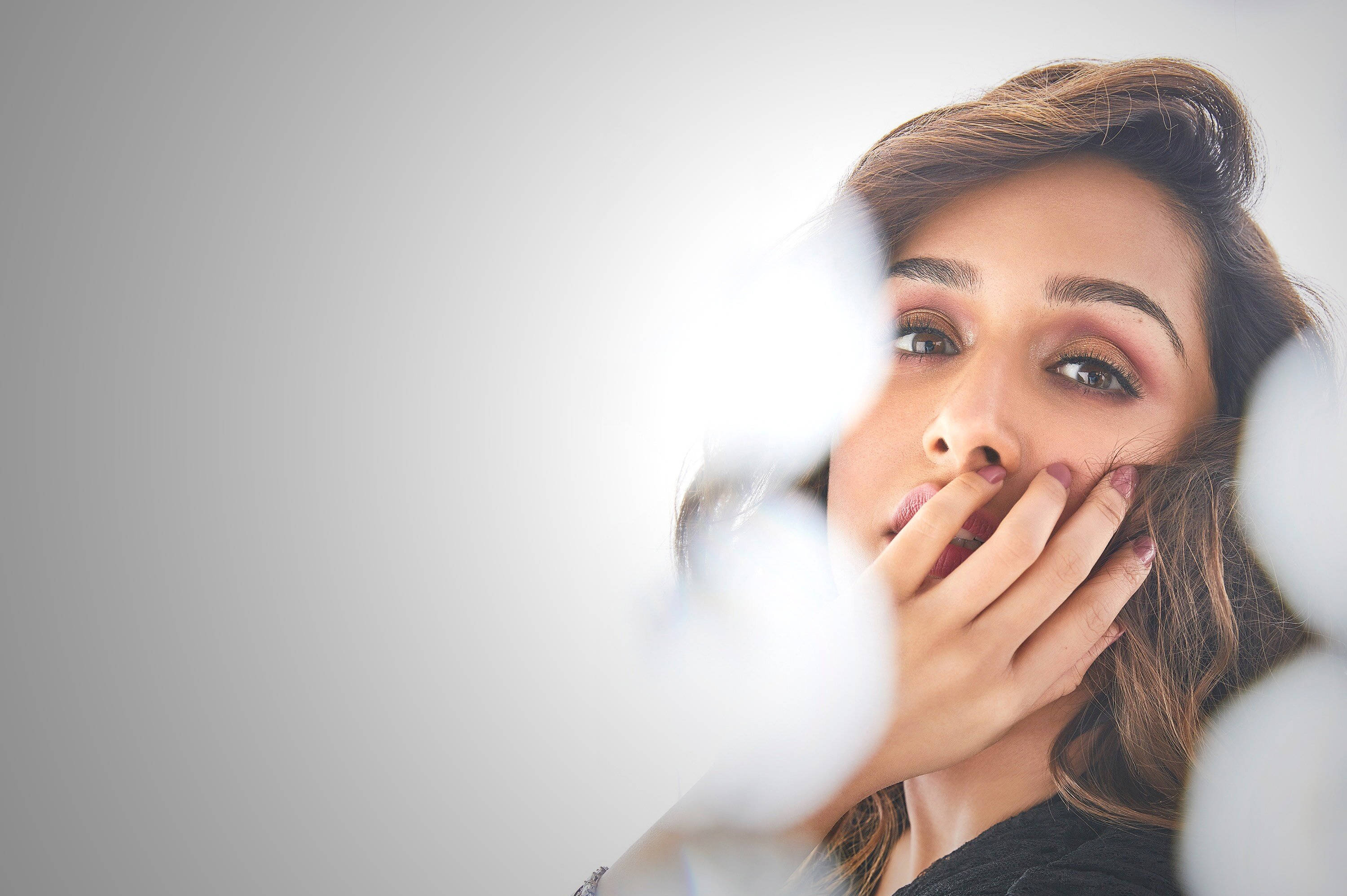 Shraddha Kapoor 桌布 壁紙 — 一位棕色頭髮、化妝的女性，在柔和的灰色背景前，透過模糊的白色球體，用手遮住嘴巴凝視。
