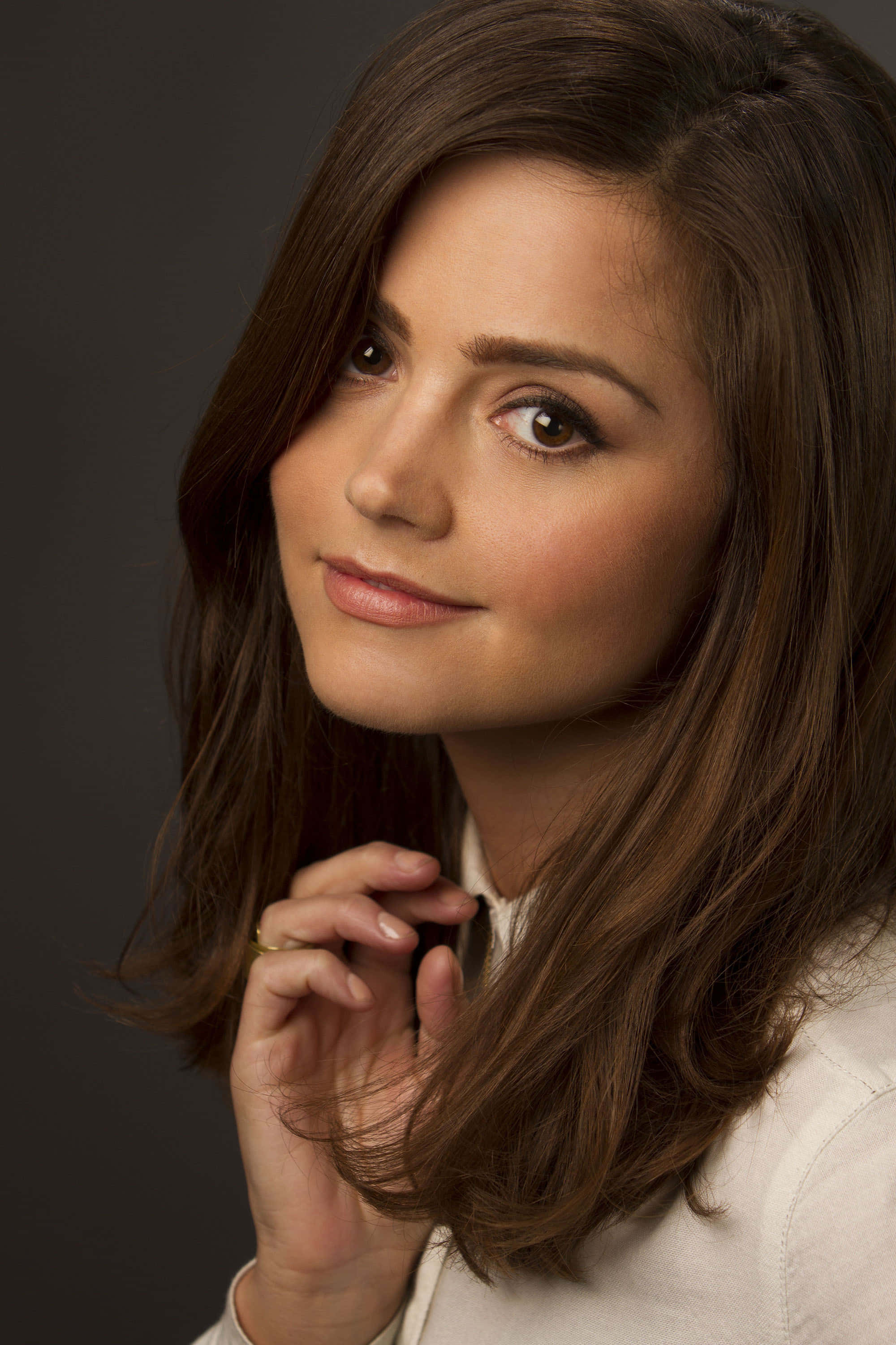 Jenna Coleman 桌布 壁紙 — 一位棕色頭髮、帶著溫柔微笑的女性，在深灰色背景前輕柔地觸摸頭髮，眼神柔和。
