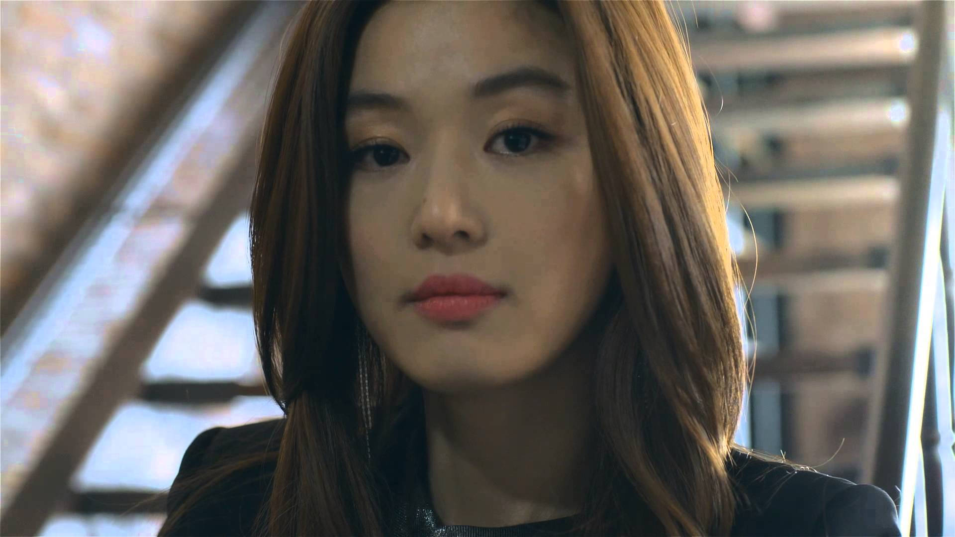 Jun Ji Hyun 桌布 壁紙 — 一位棕色頭髮、柔軟粉唇的女性，在模糊的建築背景下若有所思地凝視的特寫。