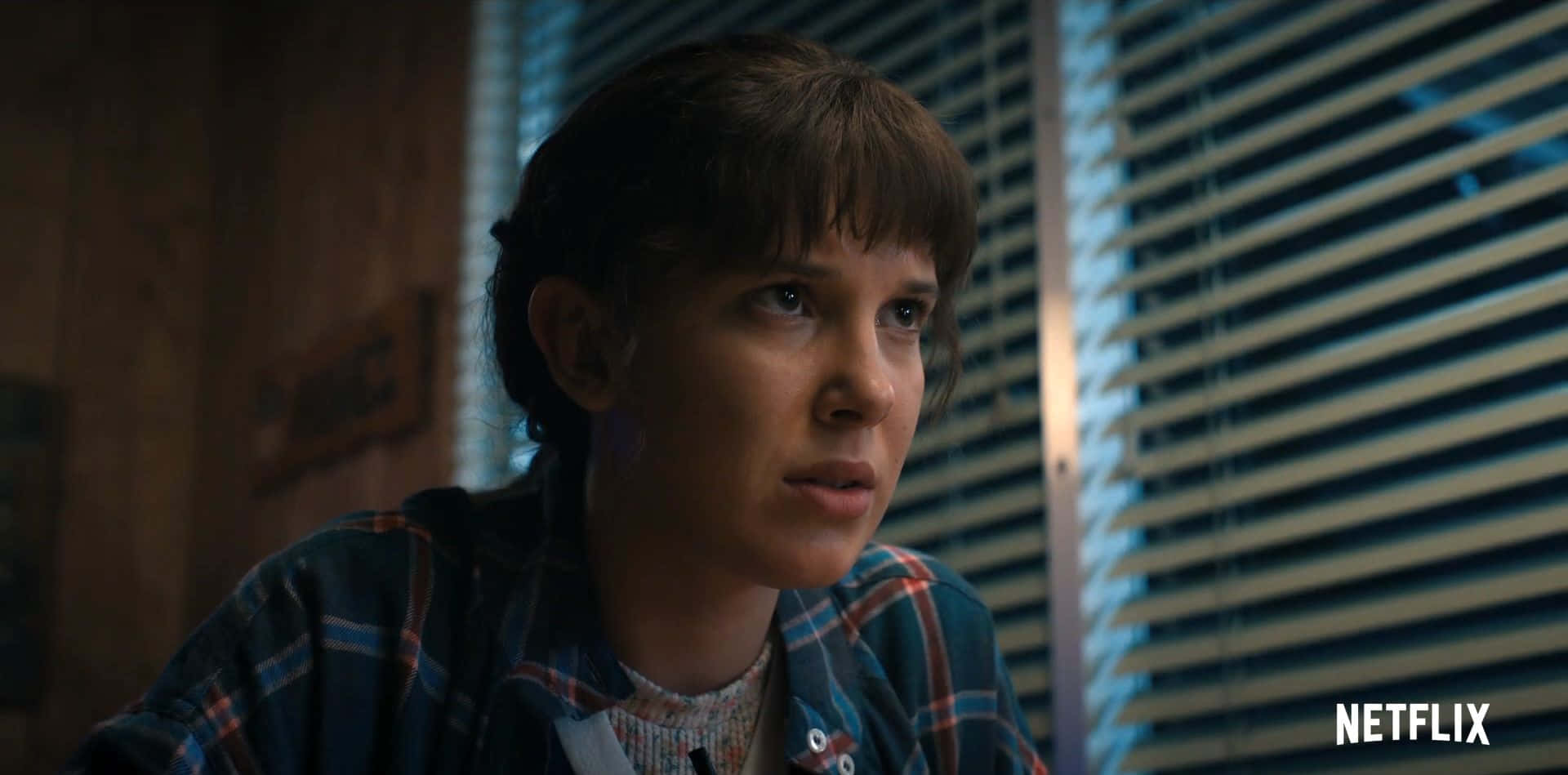 Eleven Stranger Things 桌布 壁紙 — 一位棕色頭髮、穿著格紋襯衫的年輕女性，在光線昏暗、有百葉窗的房間裡，帶著憂慮的表情看向鏡頭外。