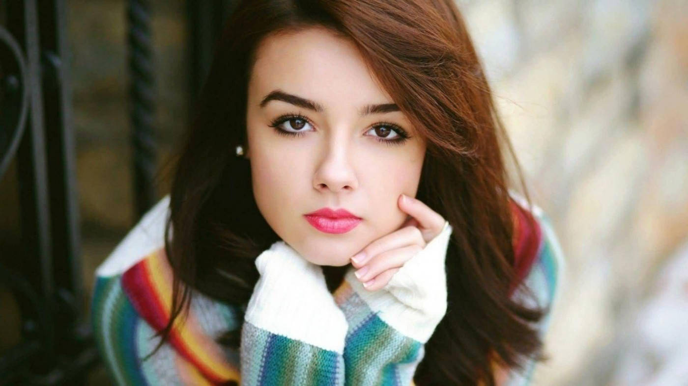 attitude 4K 桌布 壁紙 — 一位棕色頭髮、粉紅嘴唇的年輕女子，穿著色彩繽紛的條紋毛衣，將下巴擱在手上。