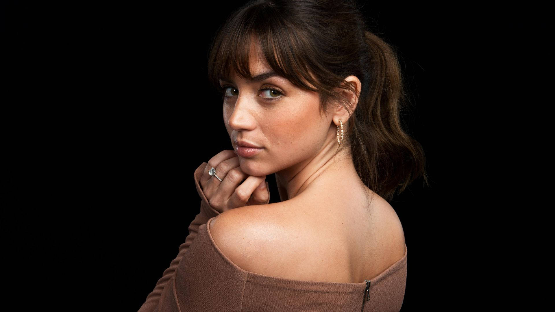 ana de armas 桌布 壁紙 — 一位棕色頭髮、綠色眼睛的女性，在黑色背景下回頭望向肩膀，穿著棕色露肩上衣。