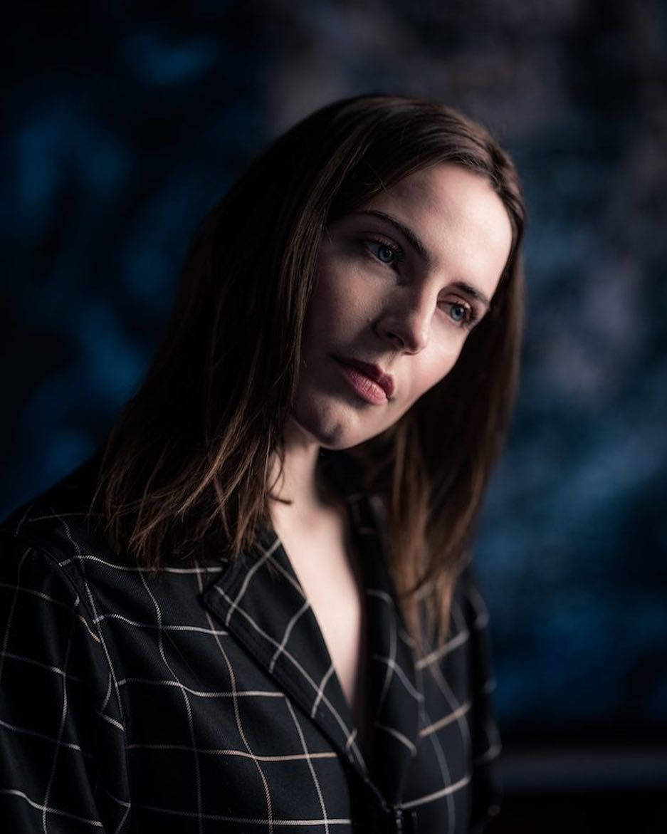 Antje Traue 桌布 壁紙 — 一位棕色頭髮、藍色眼睛的女性，身穿黑色格紋西裝外套，在深藍色工作室肖像中若有所思地凝視。