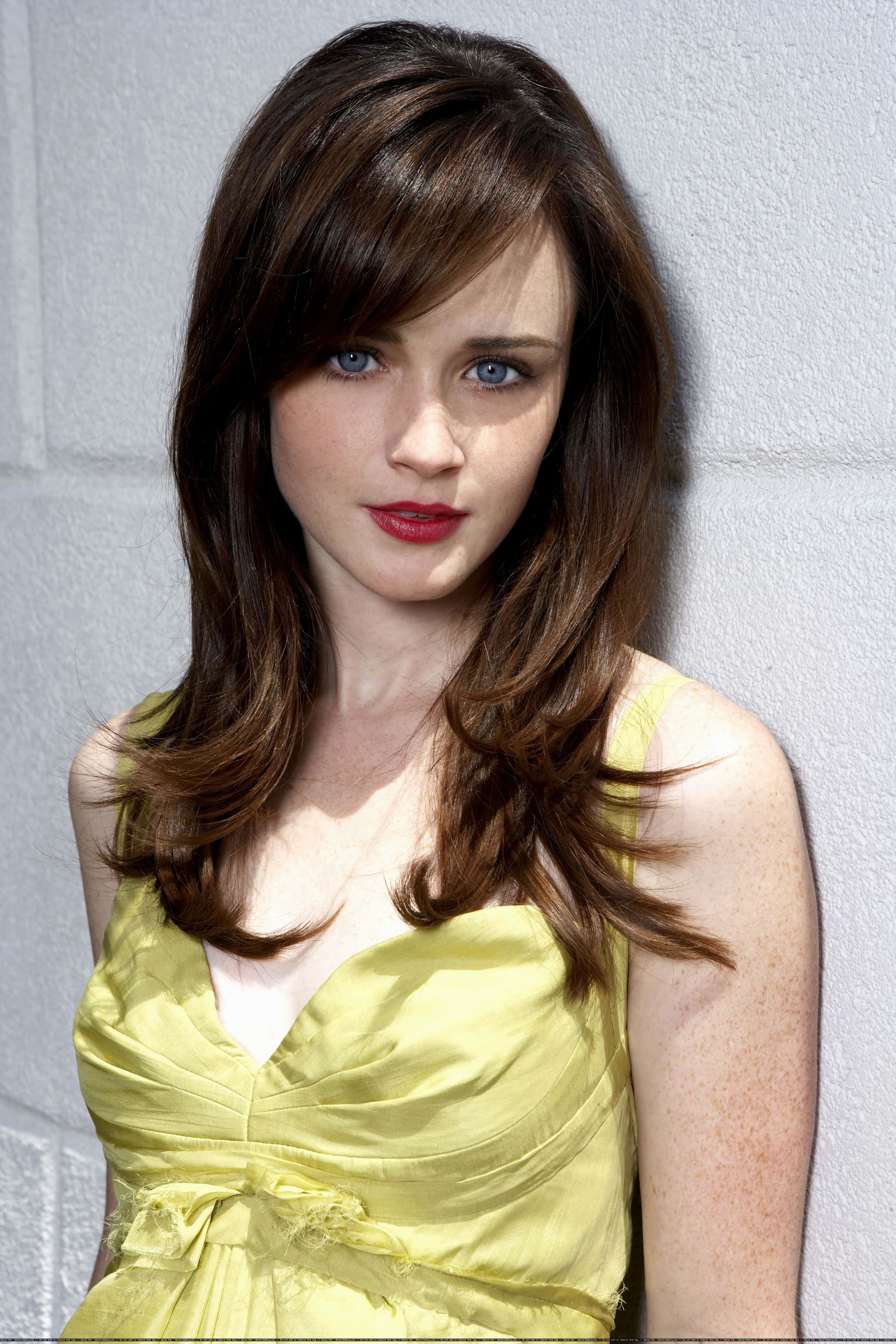 Alexis Bledel 桌布 壁紙 — 一位棕色頭髮、藍色眼睛的女性穿著黃色連衣裙，站在淺灰色磚牆前。