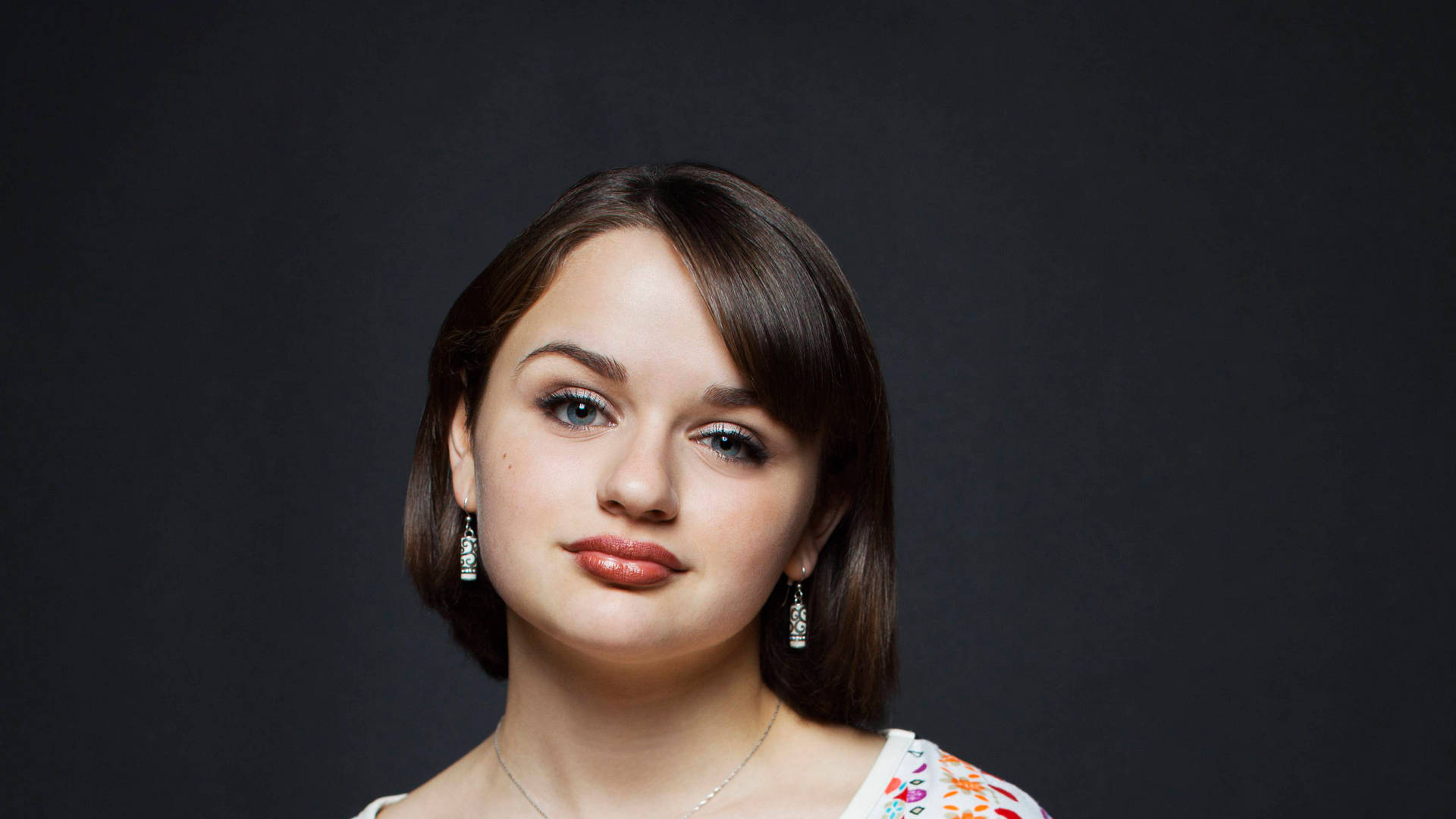 Joey King 桌布 壁紙 — 一位棕色頭髮、藍色眼睛的年輕女性在深灰色工作室背景下柔和地凝視鏡頭。
