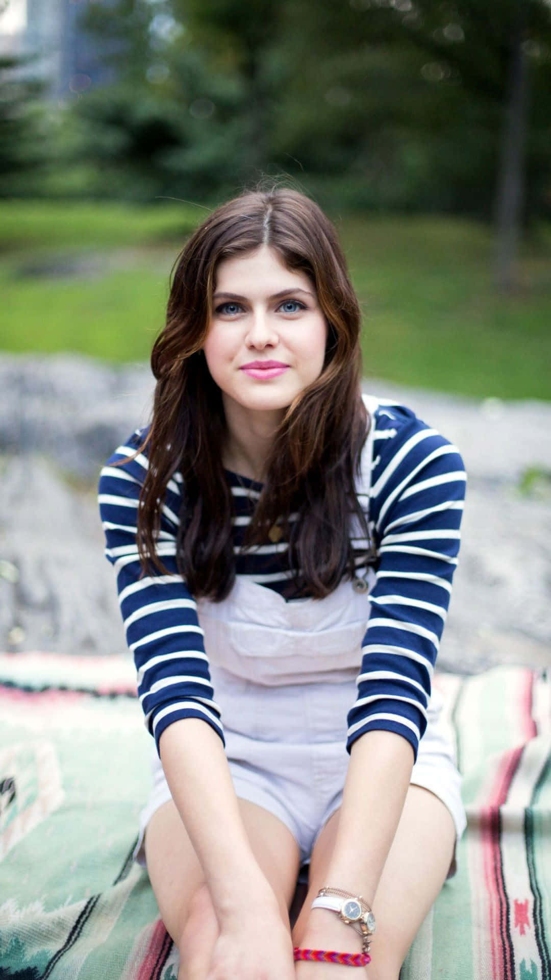 Alexandra Daddario 桌布 壁紙 — 一位棕色頭髮、藍色眼睛的年輕女性坐在戶外色彩繽紛的毯子上，穿著條紋襯衫和白色連身褲。
