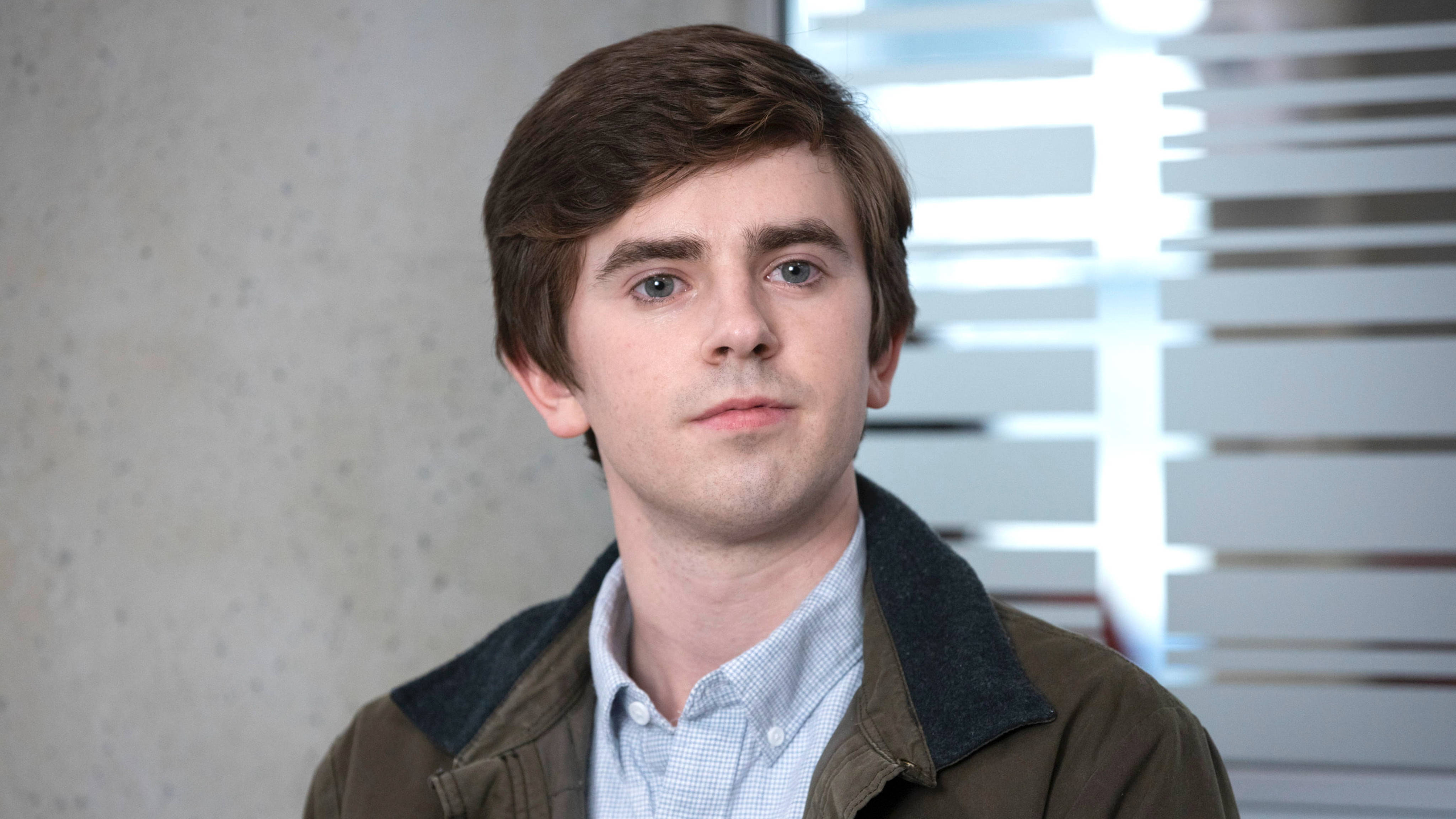 The Good Doctor 桌布 壁紙 — 一位棕色頭髮、藍色眼睛的年輕男子，身穿棕色外套內搭淺藍色襯衫，在紋理牆壁和百葉窗前直視鏡頭。